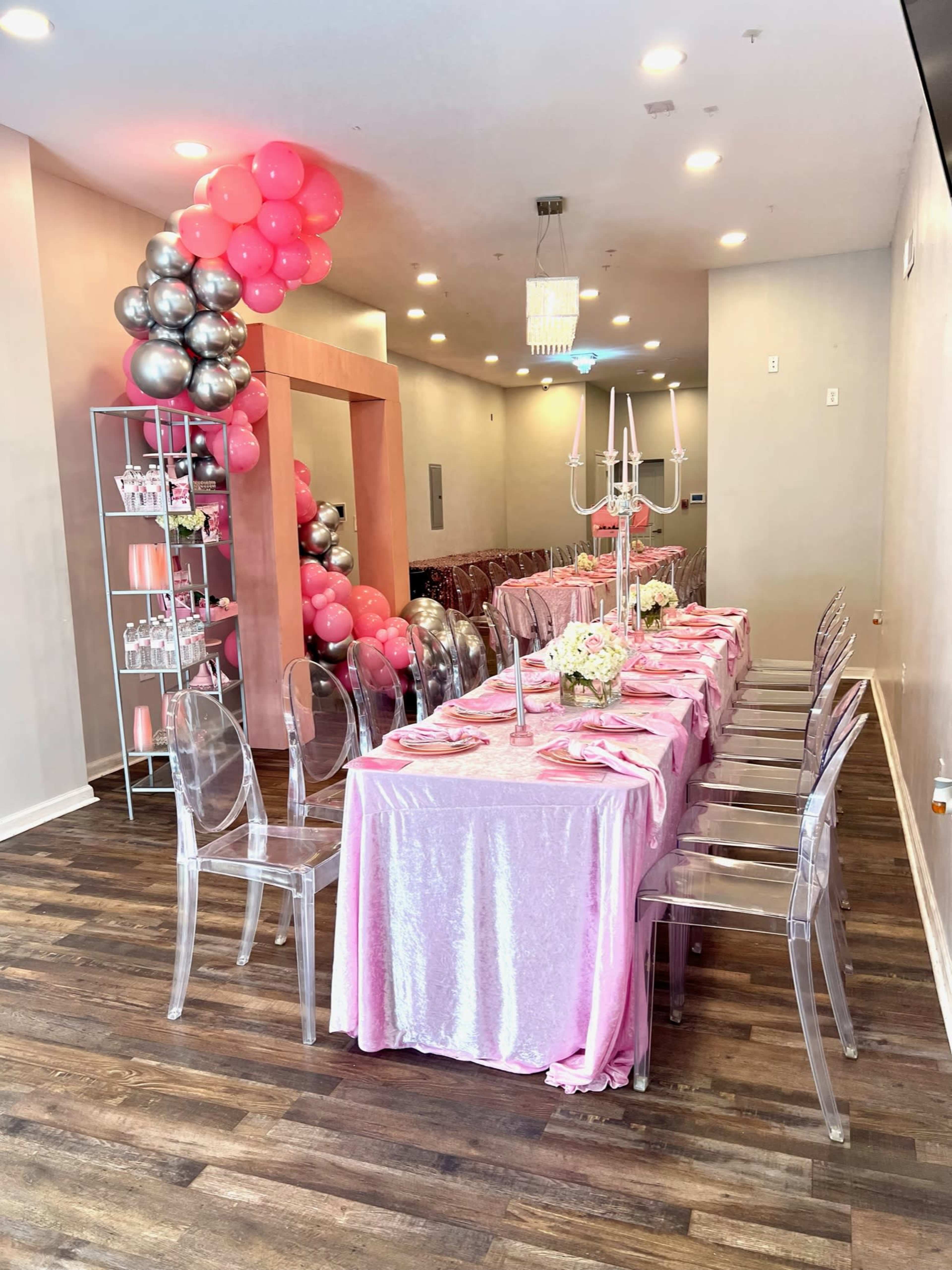 A dining area set up for an event, featuring a long pink table with chairs, adorned with a floral centerpiece and balloons.