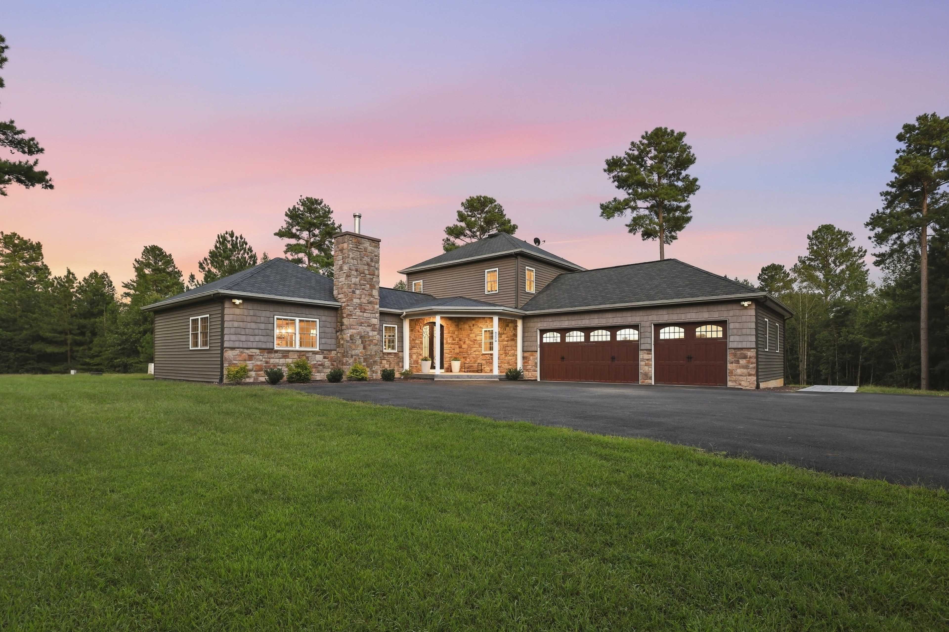 The image shows a modern two-story house with a stone chimney, surrounded by green grass and tall trees under a colorful sky at dusk.