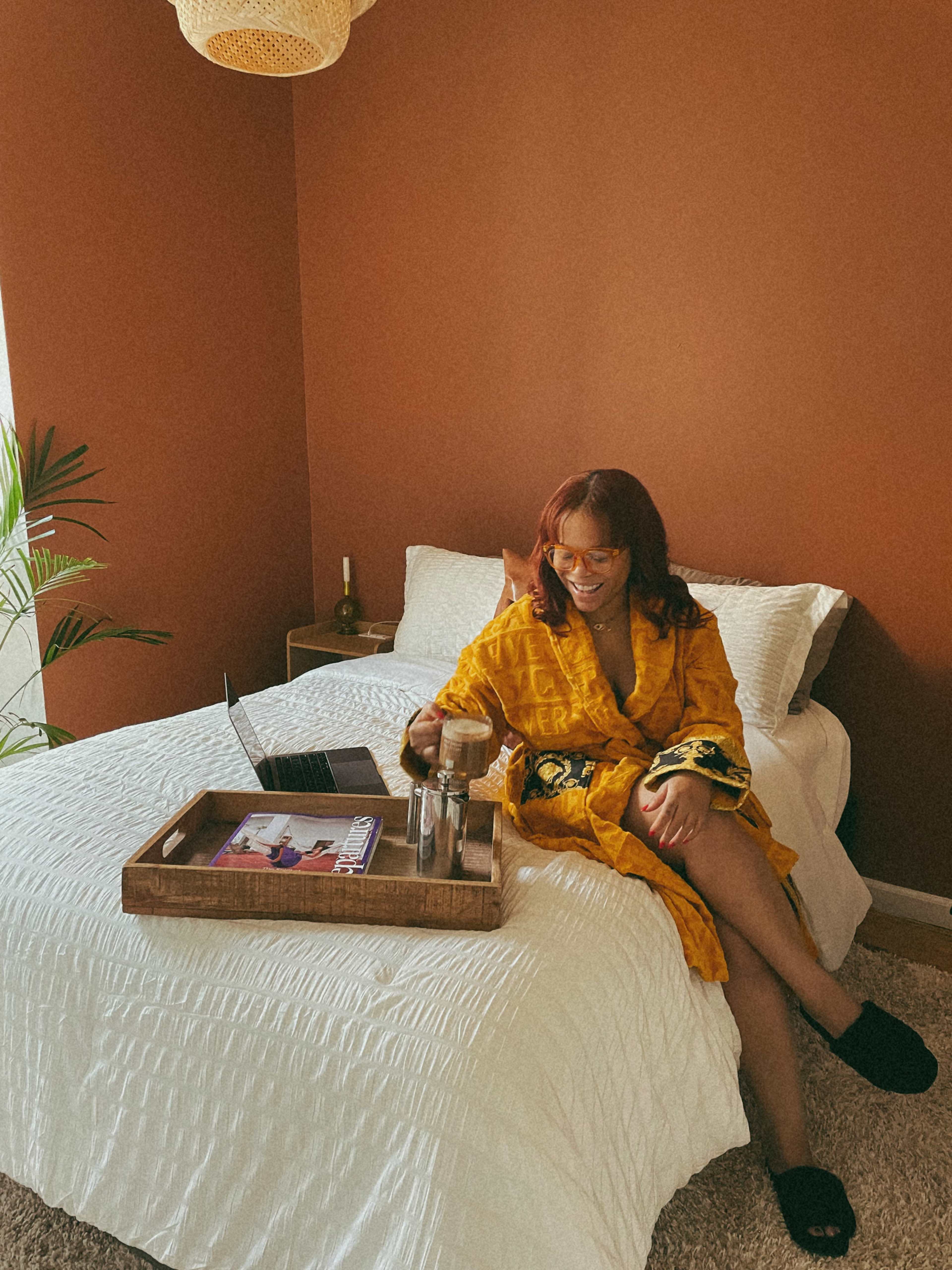 A woman in a yellow robe sits on a bed with a laptop and a tray of drinks in a room with brown walls.