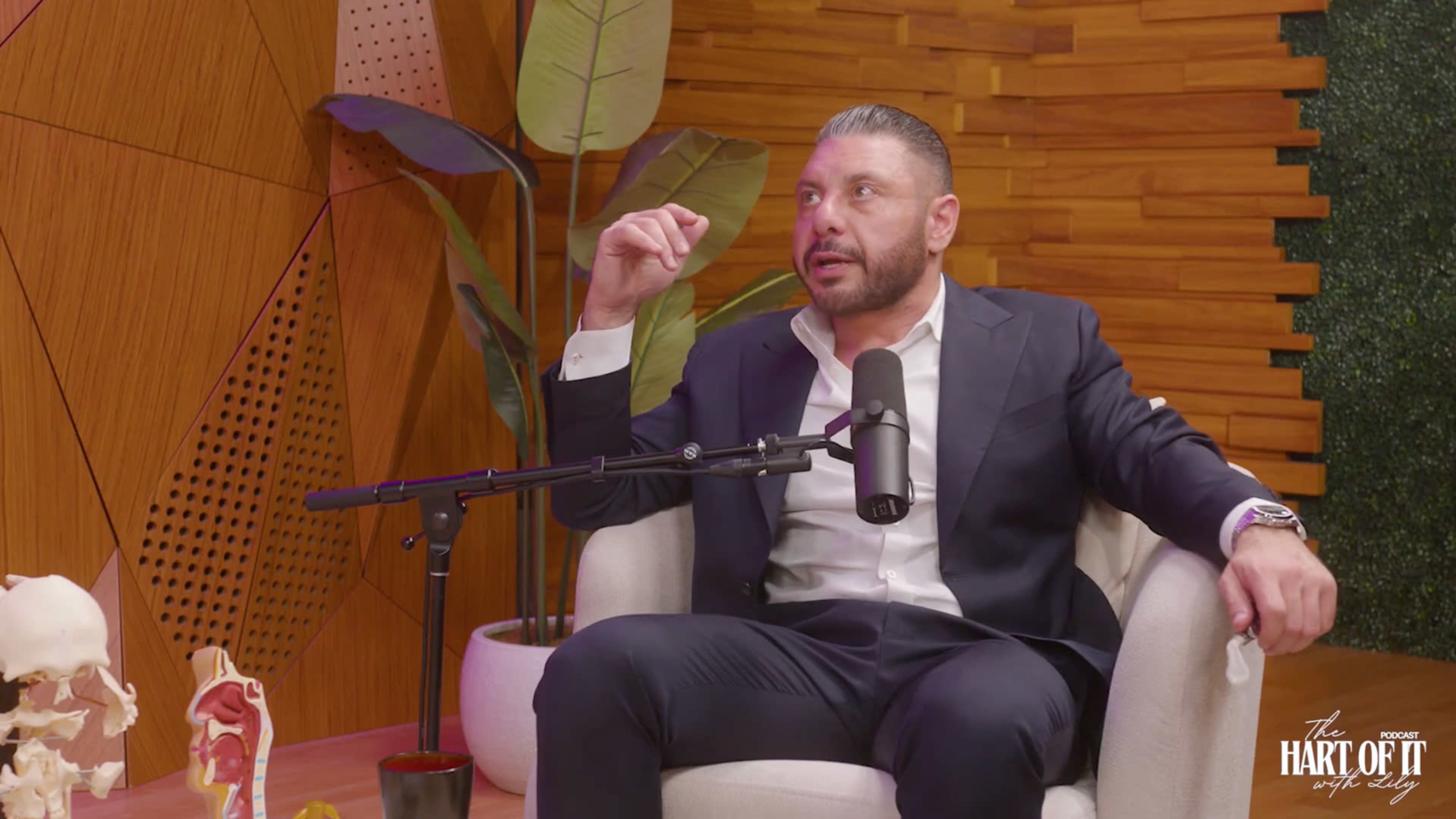 A man in a suit sits in a chair with a microphone in front of him, surrounded by wooden paneling and decorative plants.