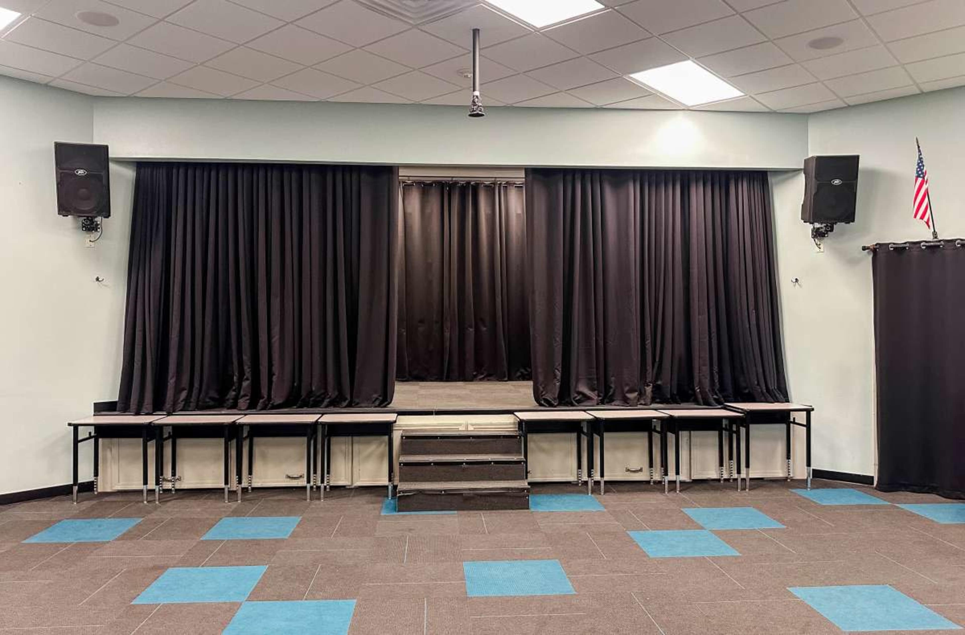 The image shows a stage with black curtains and speaker systems on either side, flanked by several tables in front, all set in a room with blue and gray tiled flooring.