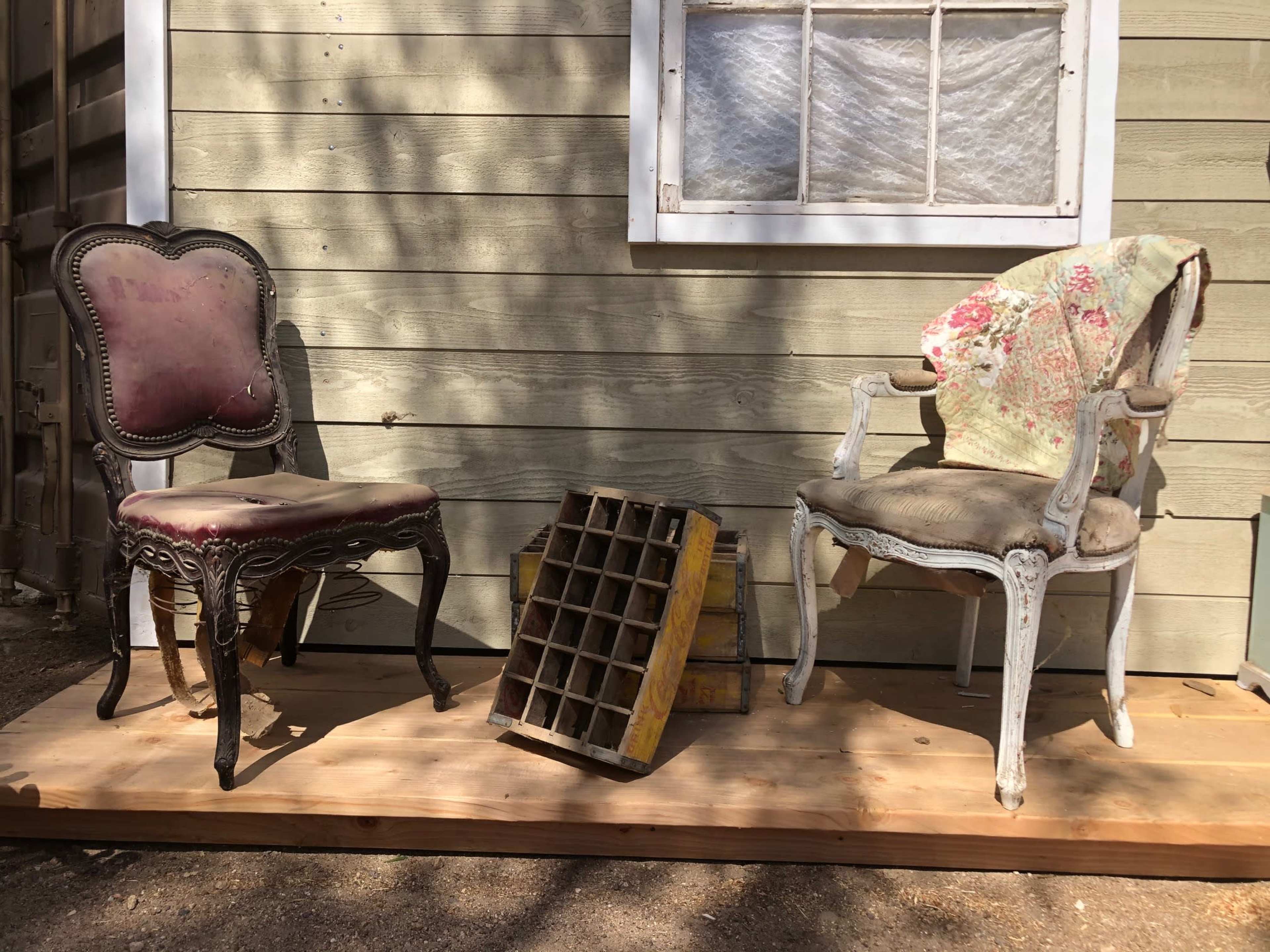 Two vintage chairs, one upholstered in purple and the other white with a floral blanket draped over it, are positioned beside a wooden crate and a divided wooden tray on a porch.