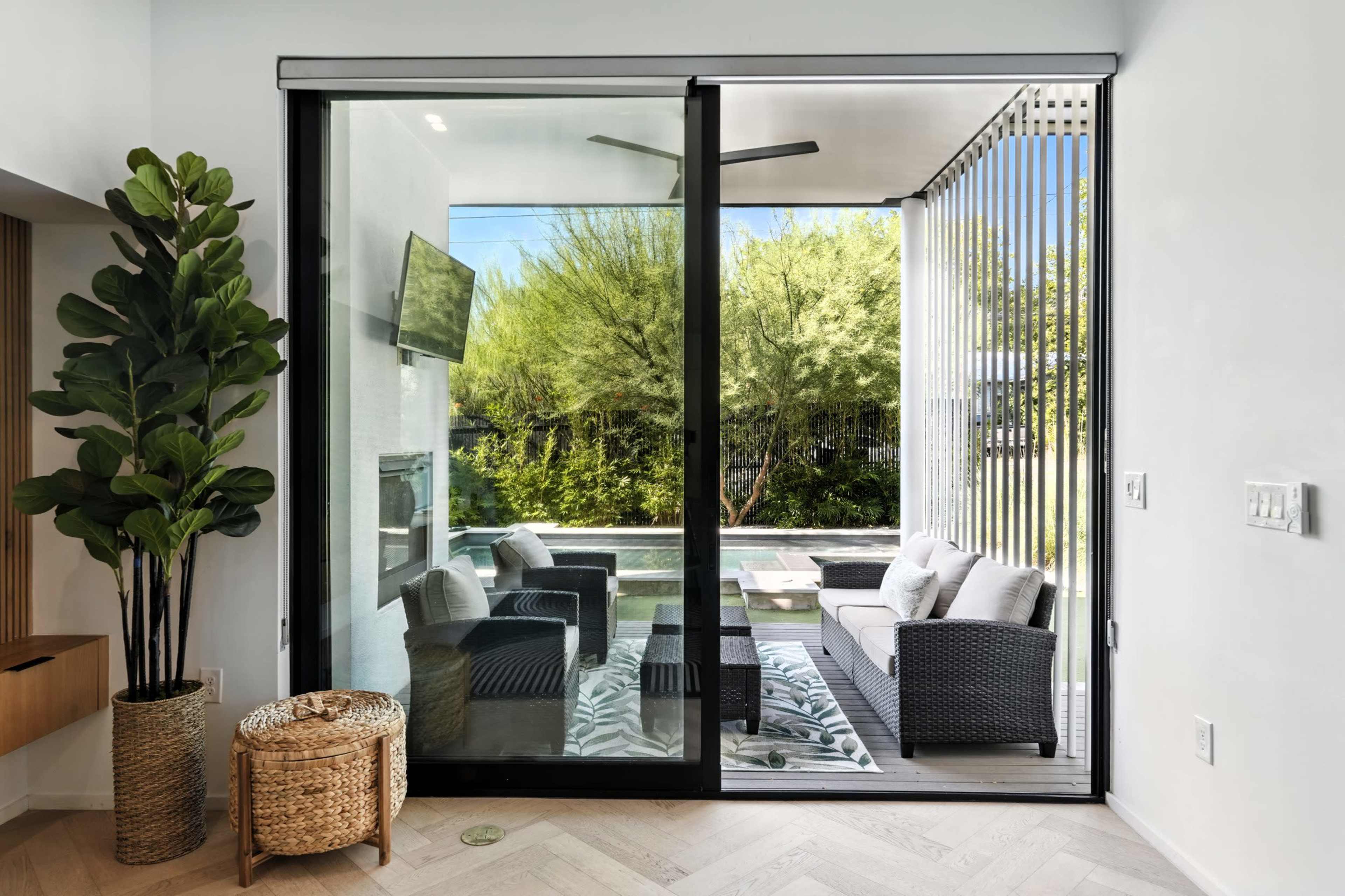 A modern living space features large glass doors opening to a patio with outdoor seating and greenery visible beyond.