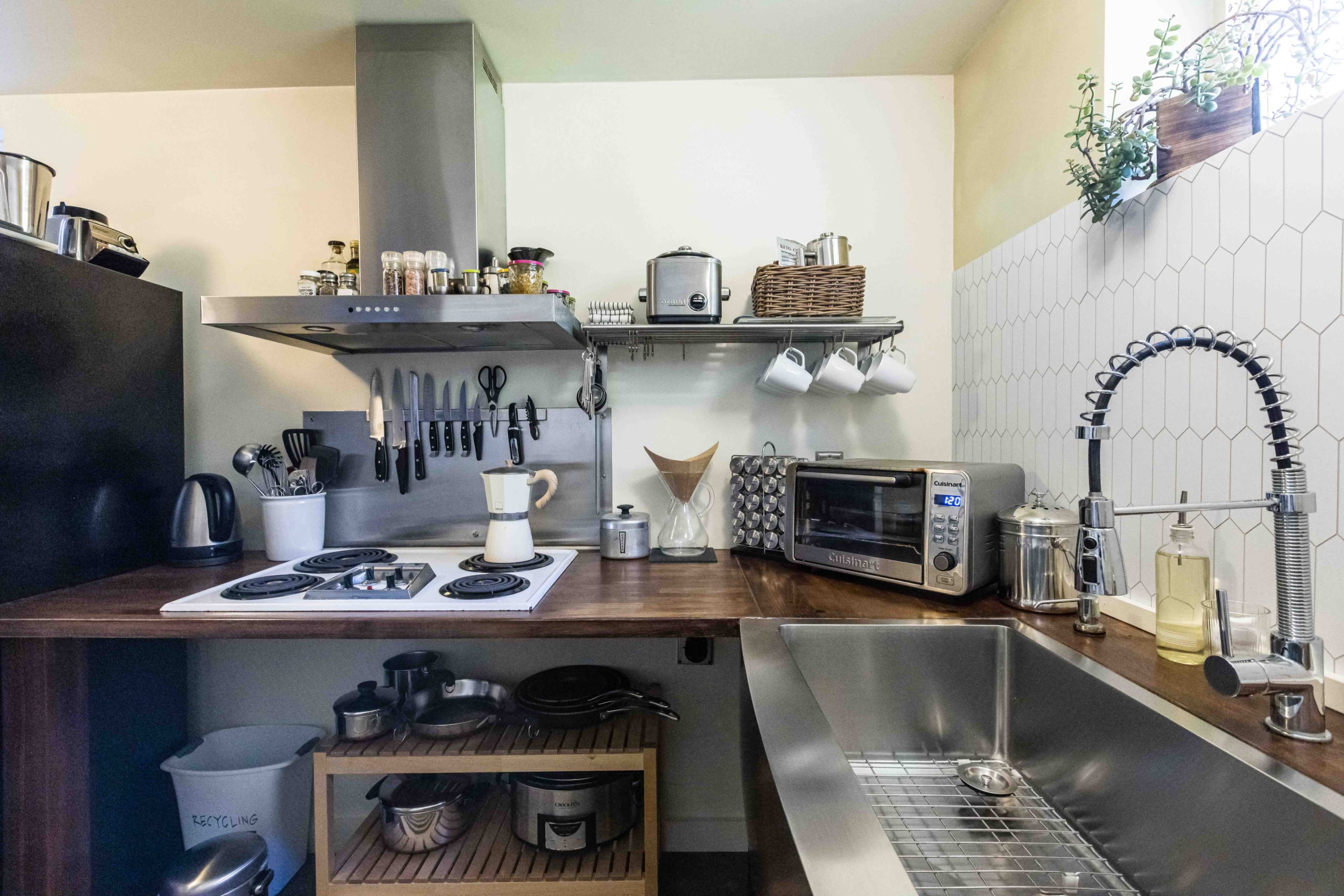 The image shows a modern kitchen featuring a wooden countertop with a stovetop, an oven, and kitchen utensils mounted on a wall, alongside a stainless steel sink and various kitchen appliances.