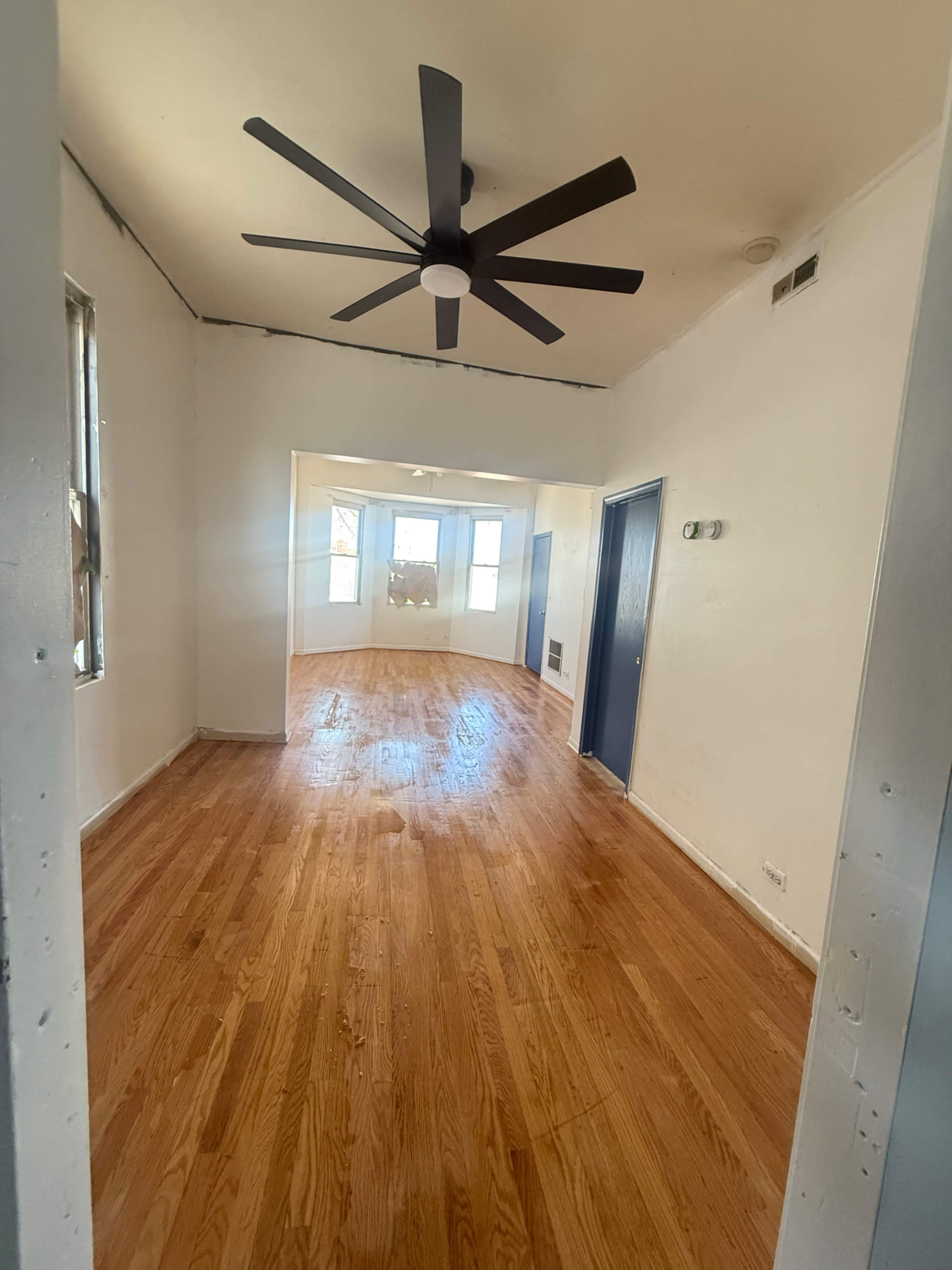 The image shows a room with hardwood floors, a ceiling fan, and windows that allow natural light to enter, connecting to another space in the background.