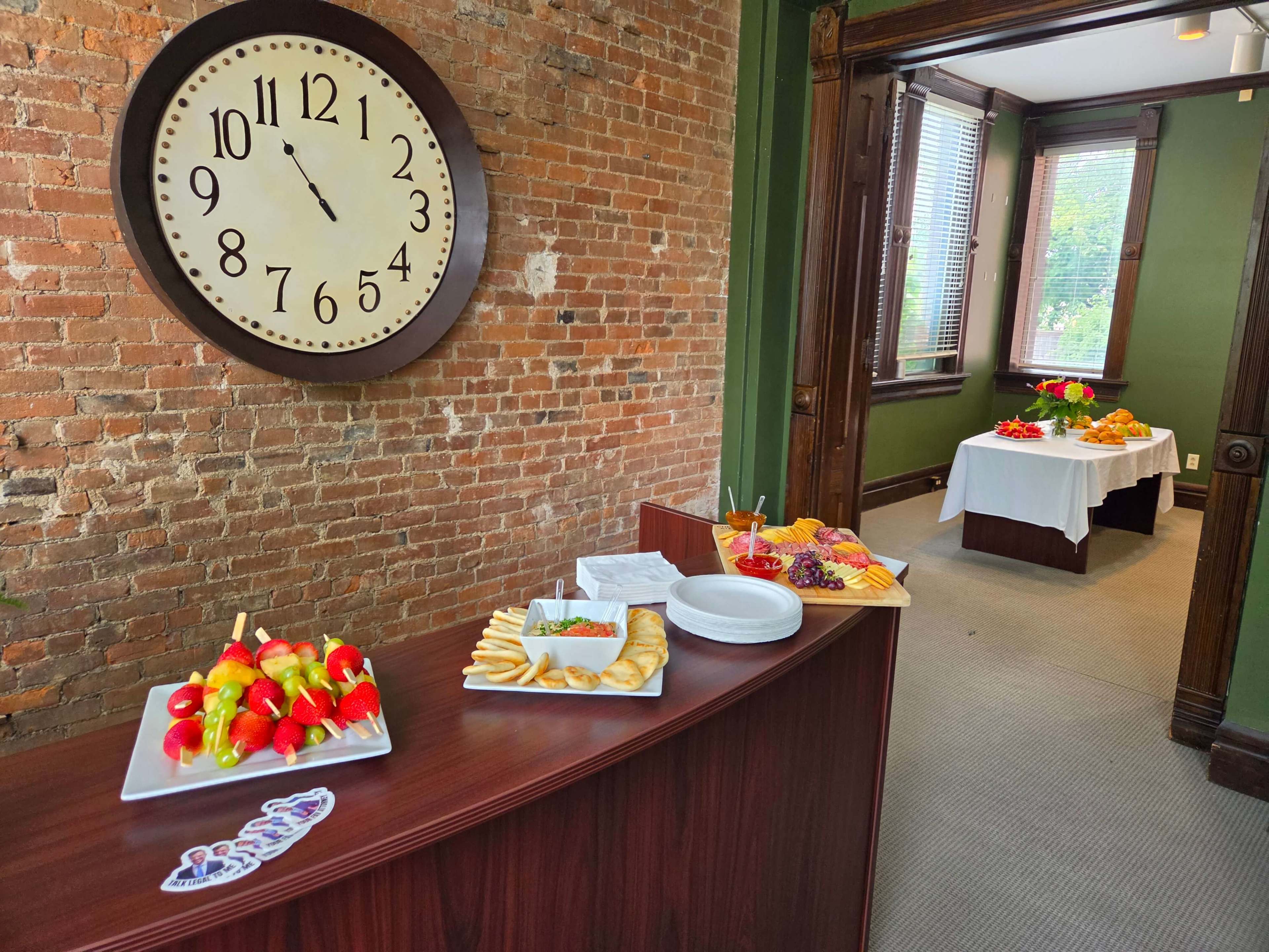 The image shows a room with a wooden counter displaying various food platters, a large clock on the brick wall, and two tables set with additional food in the background.