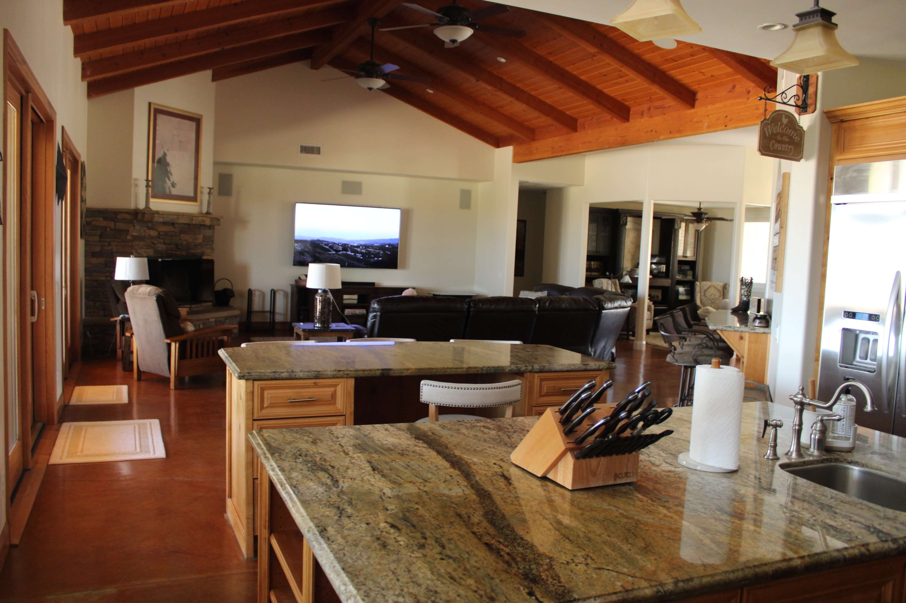 The image shows a spacious kitchen and living area with wooden ceiling beams, a large island with a granite countertop, and a wall-mounted television in the background.