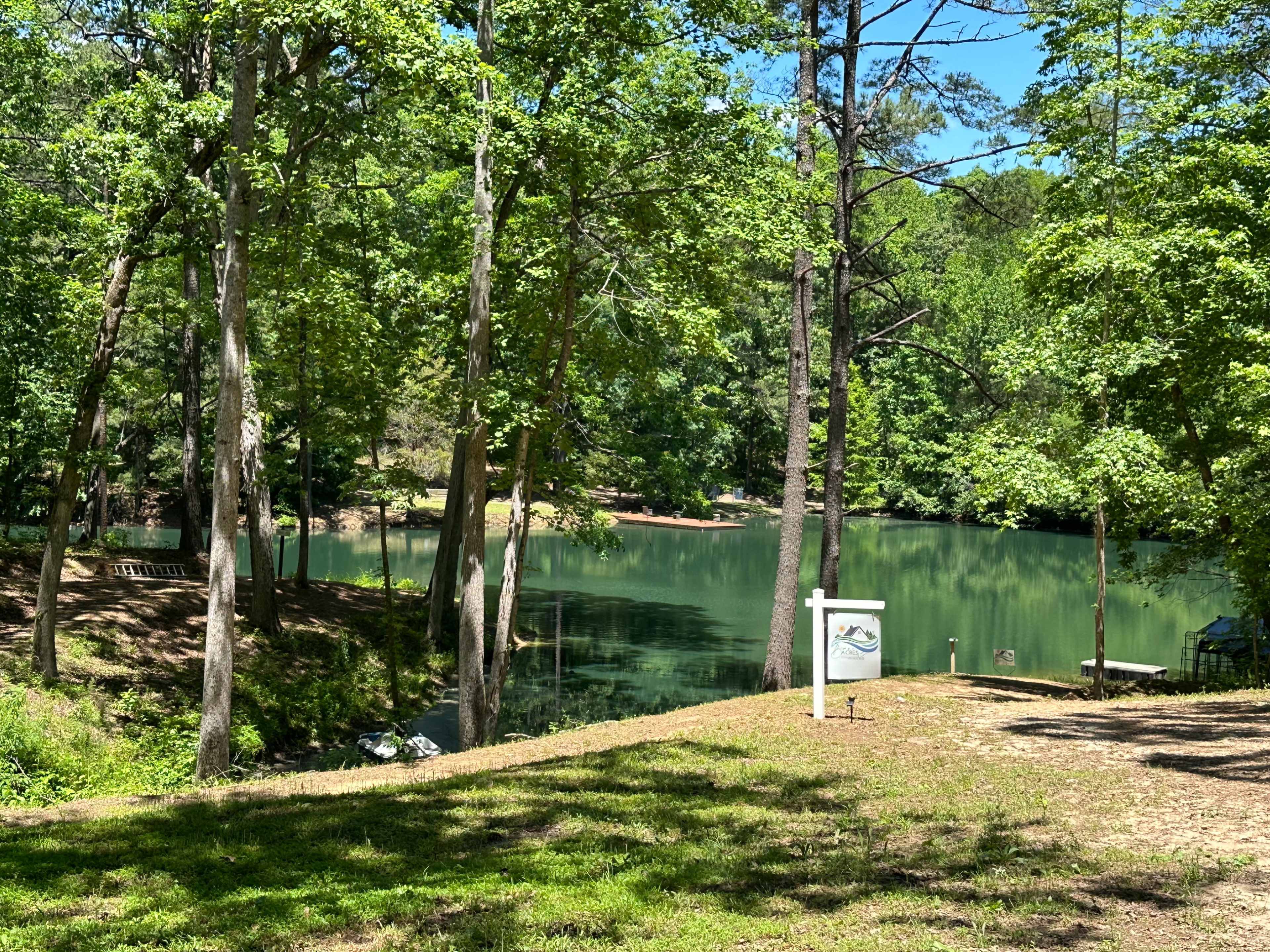 A tranquil lake surrounded by tall trees and a grassy area, with a signpost near the water's edge.