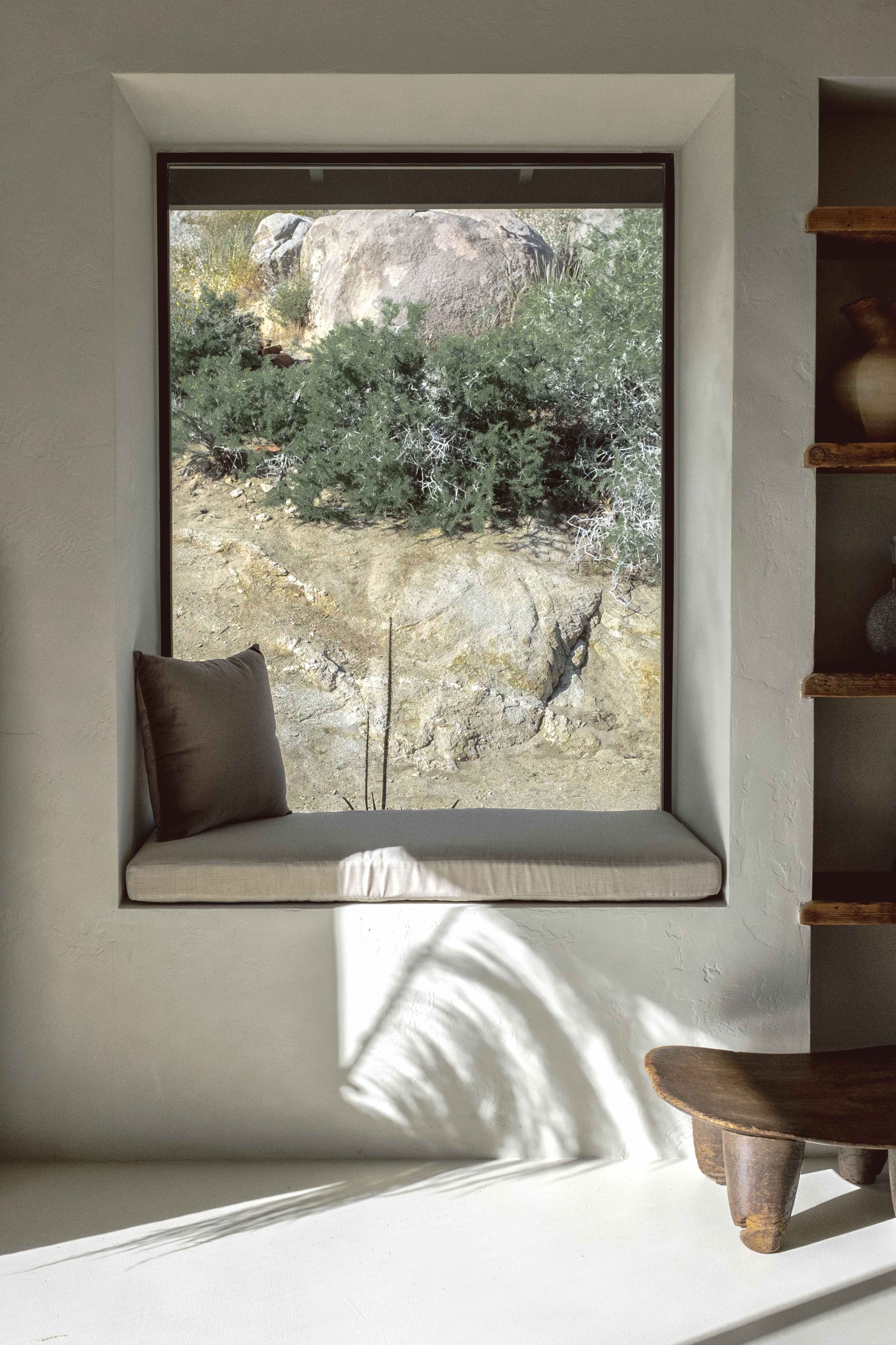 A window with a cushioned seat overlooks a rocky landscape with dense greenery.