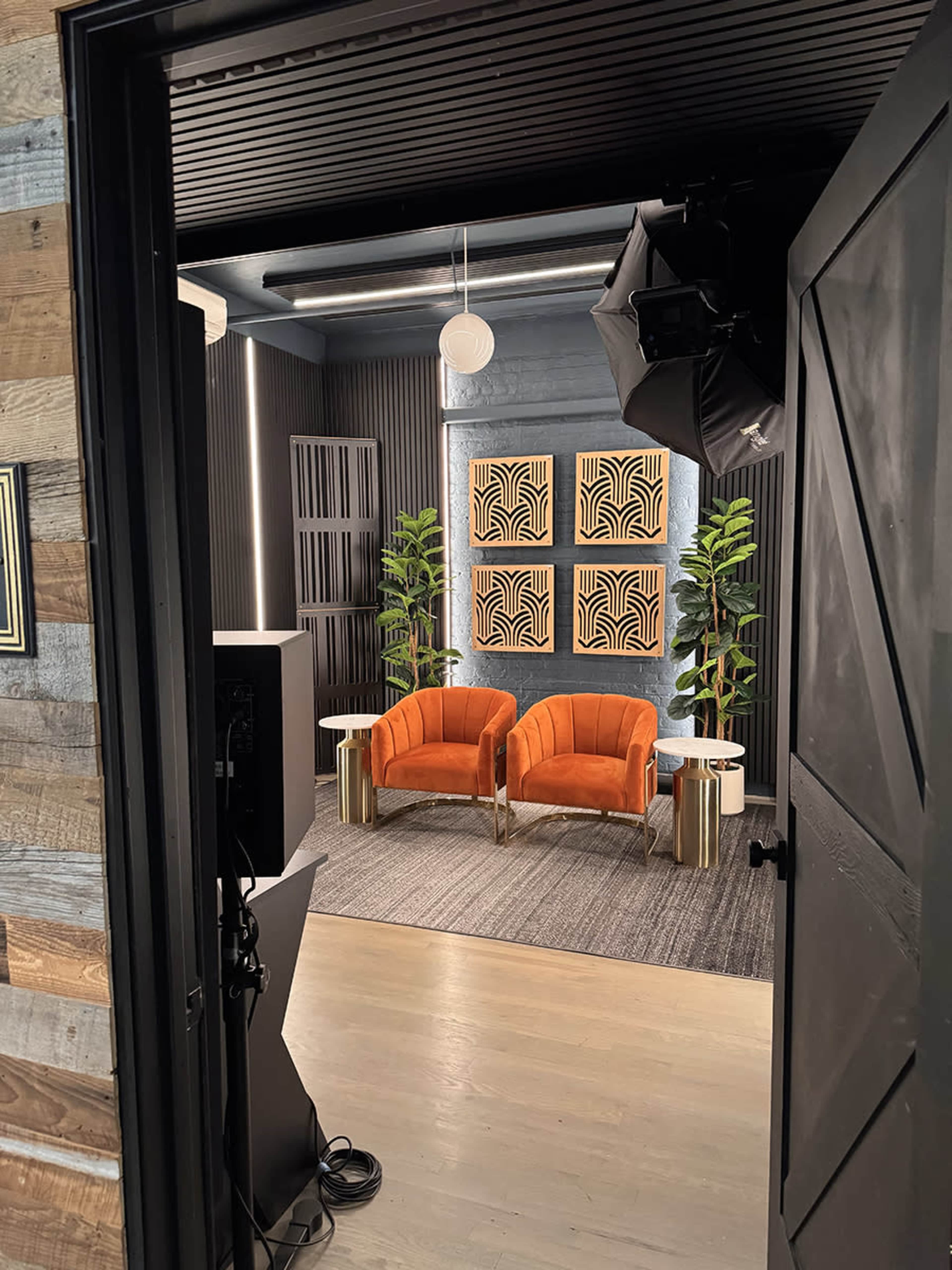 The image shows a well-lit studio space featuring two orange chairs, two decorative wall art pieces, and potted plants arranged around a small table.
