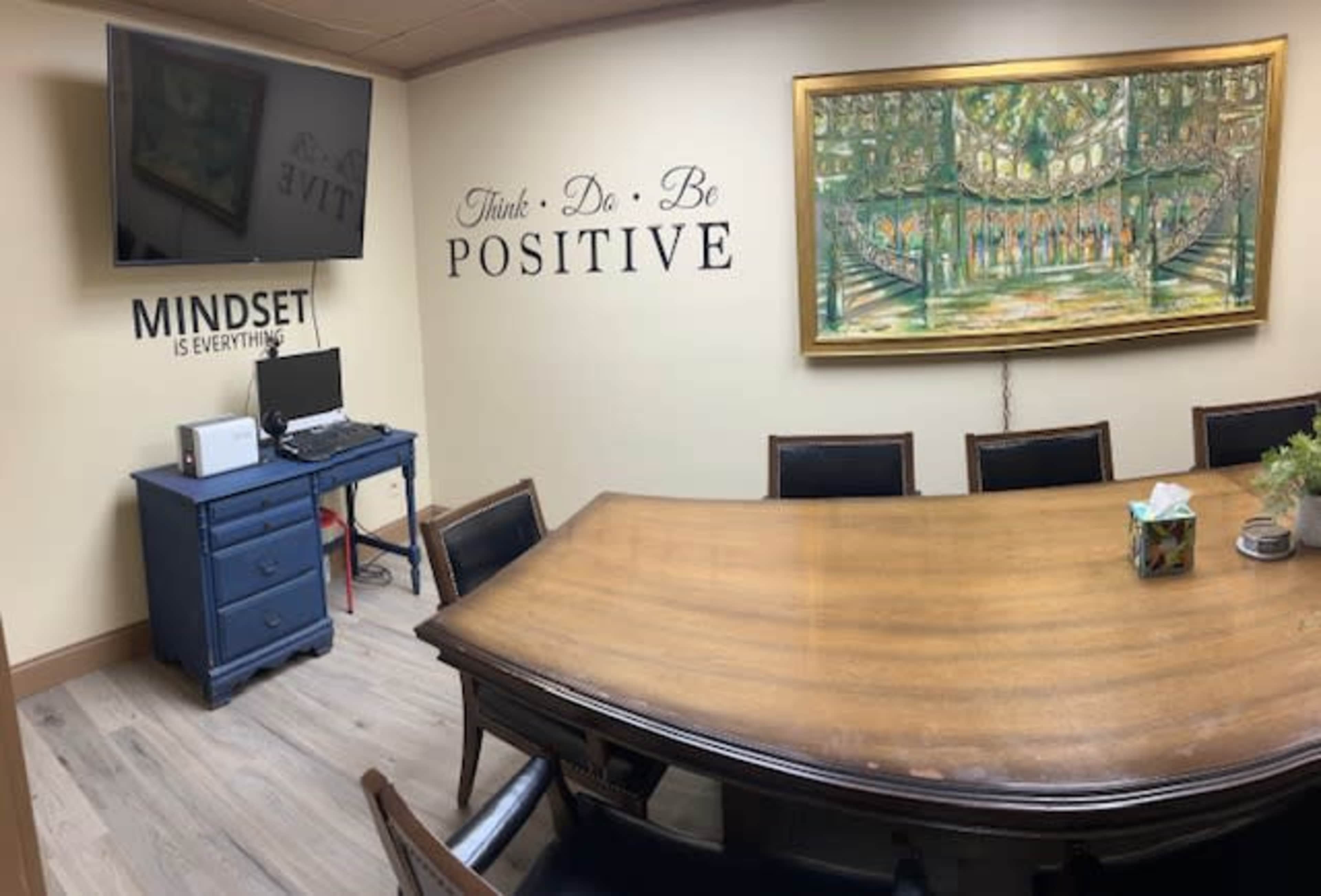 A meeting room with a large wooden table, a blue cabinet with a monitor on it, and motivational phrases painted on the wall alongside a decorative painting.