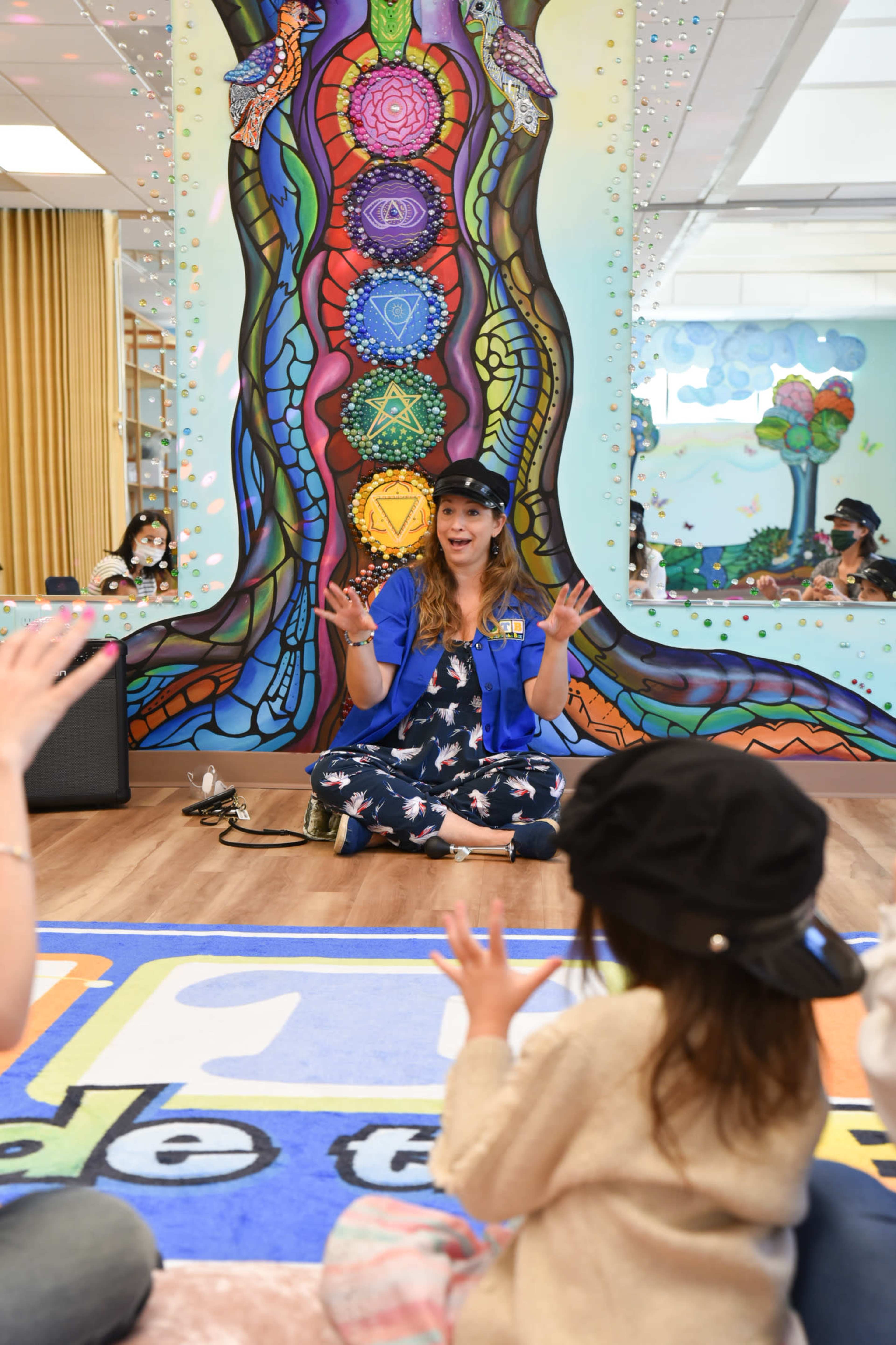 A woman in a blue jacket sits cross-legged on the floor in front of a colorful mural, interacting with an audience of children.