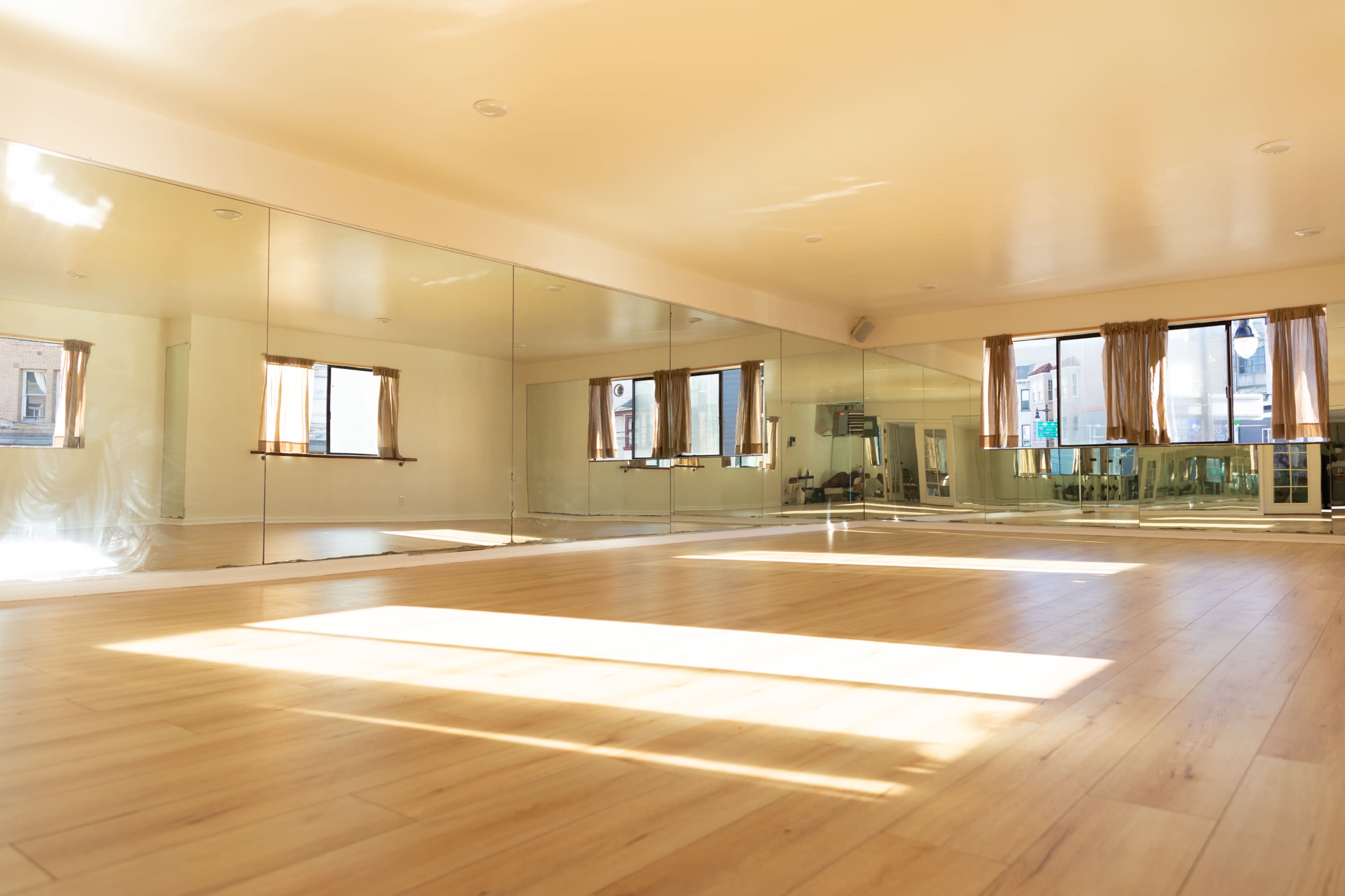 A spacious room with wooden flooring and large mirrors along one wall, illuminated by sunlight coming through windows with curtains.