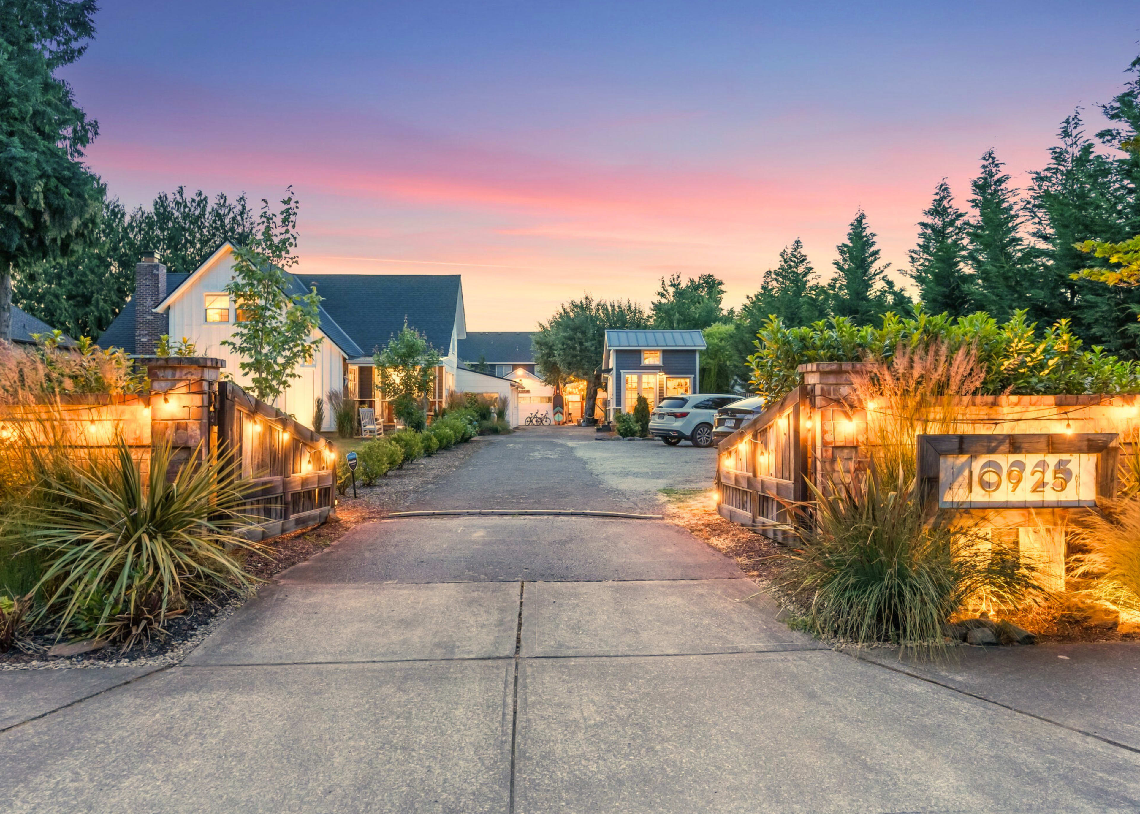 A well-lit driveway leads to a house surrounded by trees and landscaping at sunset.