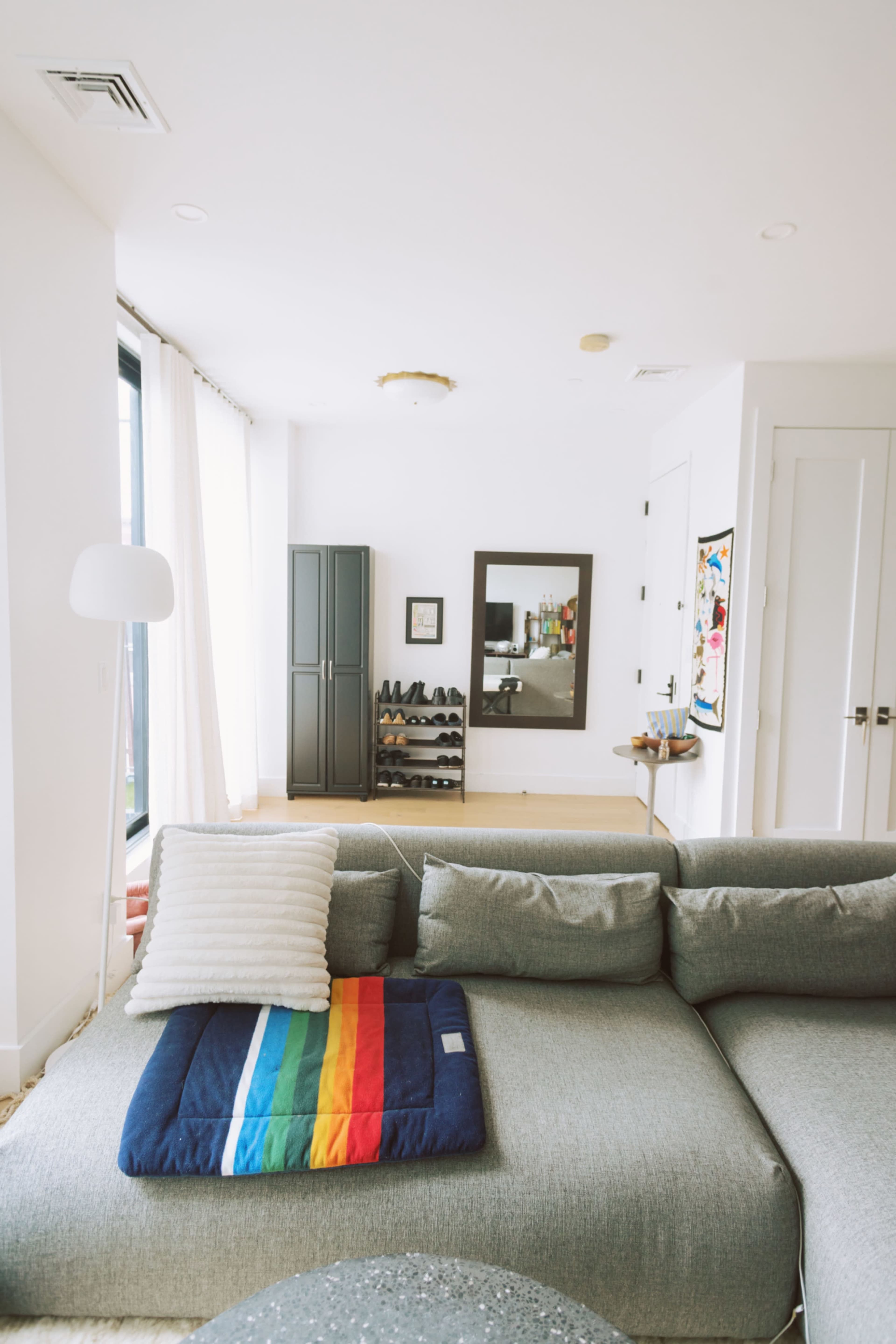 The image shows a modern living room with a gray sectional sofa, a rainbow-striped blanket, a floor lamp, and a mirror reflecting a shelf with shoes.