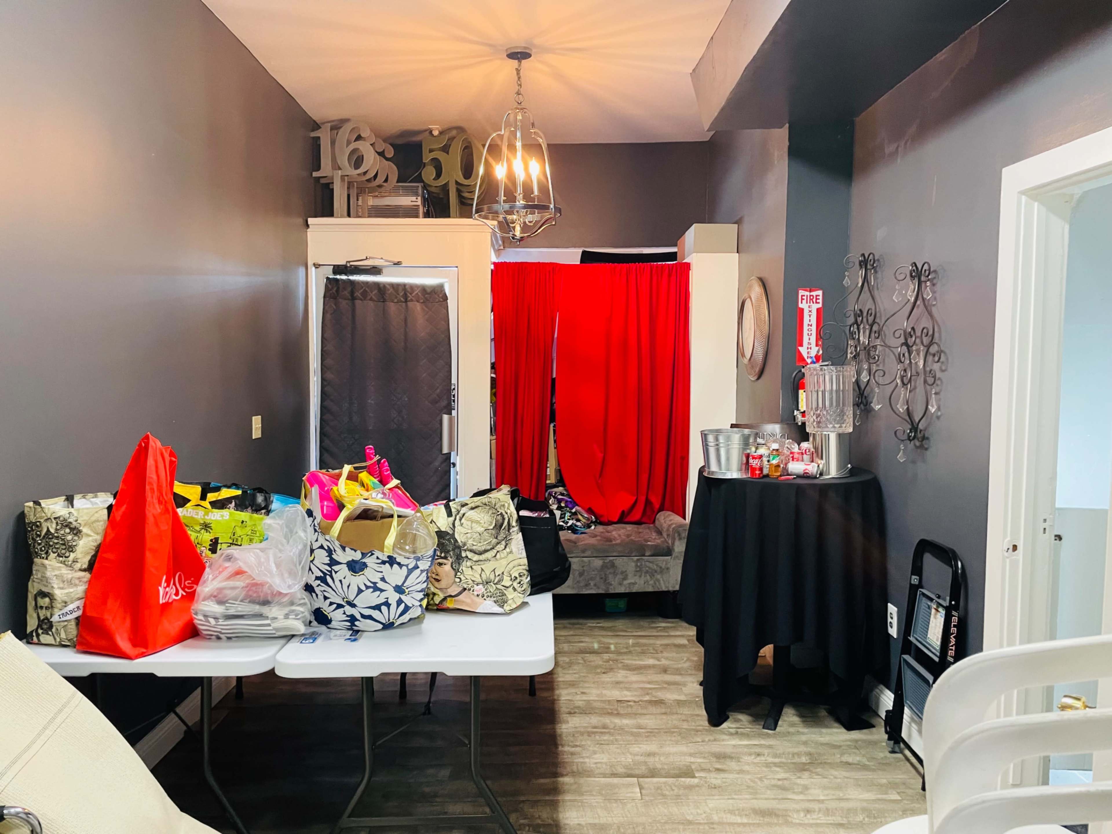 A cluttered room contains bags and a table, with a red curtain at the back and a black table set up nearby.