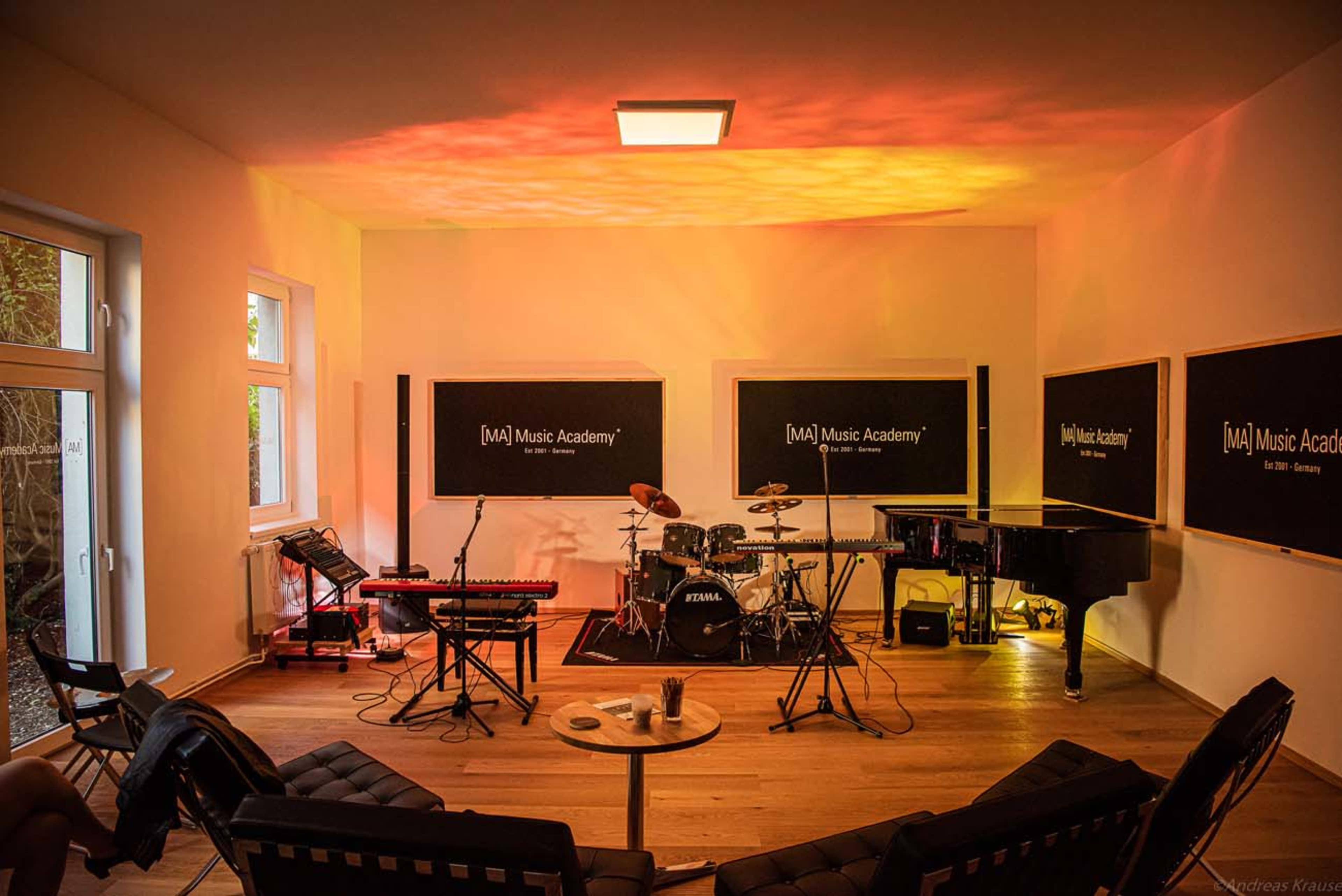 The image shows a spacious music studio equipped with a piano, drum set, and keyboard, surrounded by chairs arranged in a semi-circle.