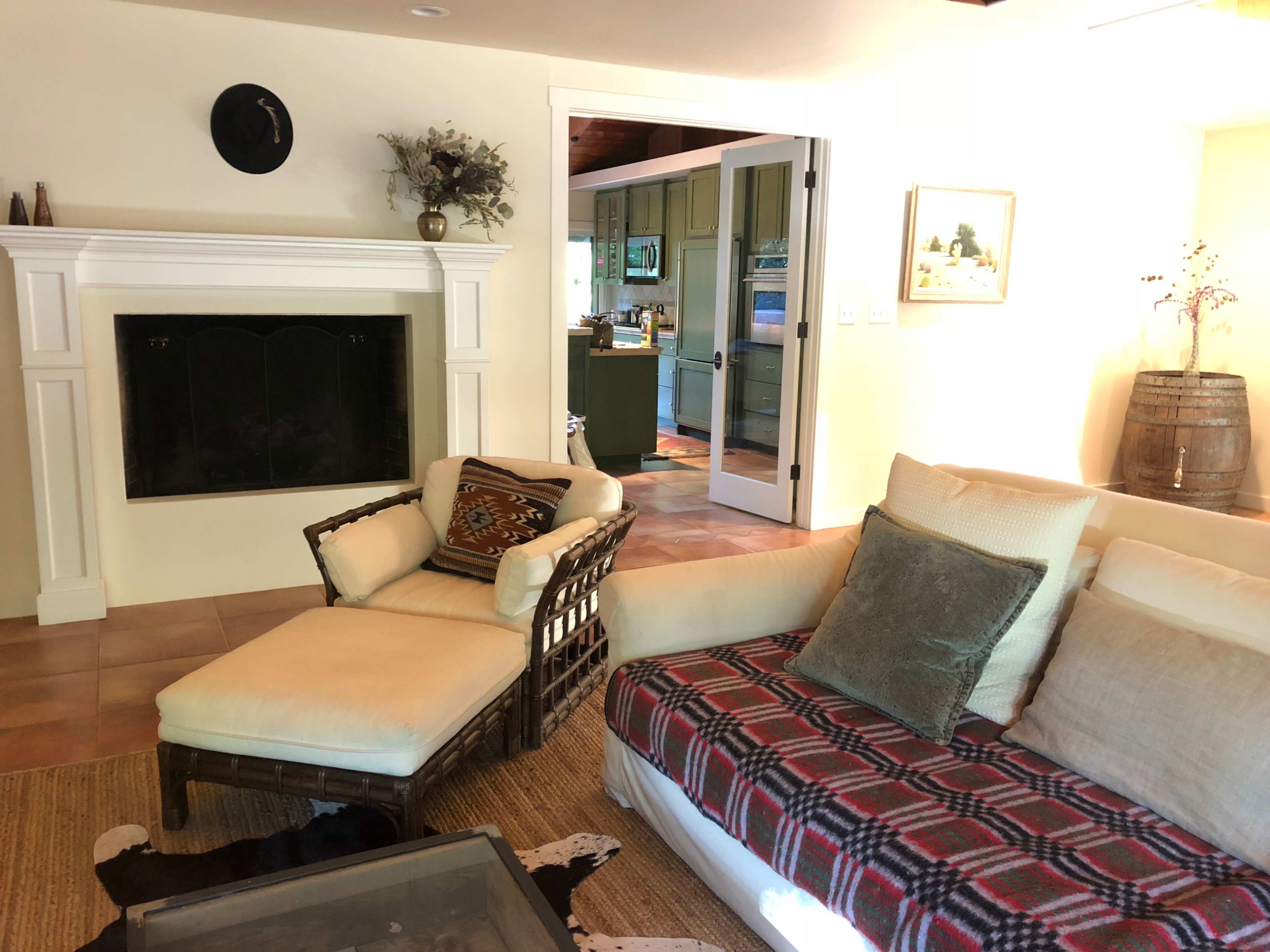 A living room with a fireplace, a wicker chair, a plush couch covered with a plaid blanket, and a doorway leading to a kitchen in the background.