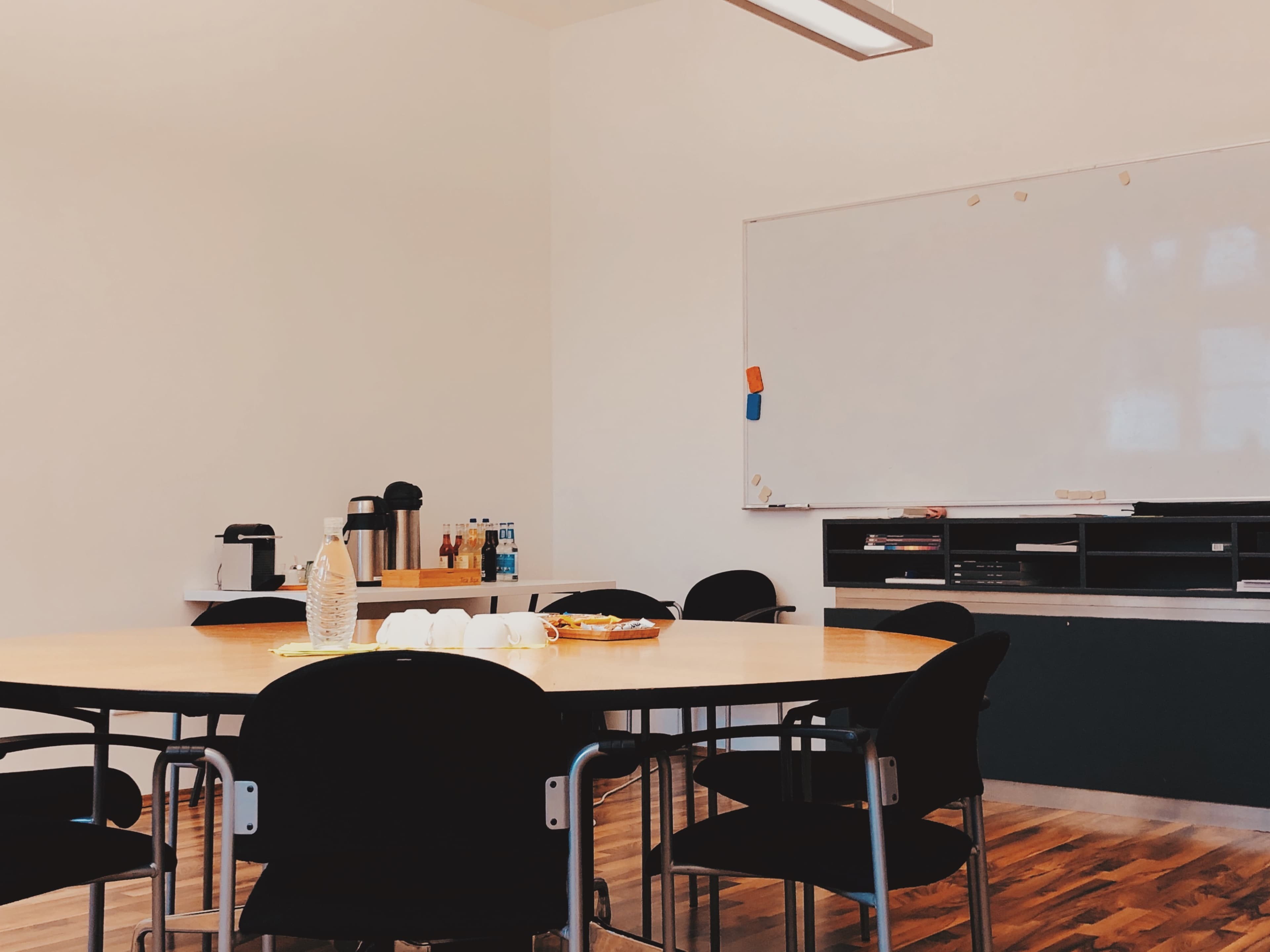 A conference room features a large round table surrounded by black chairs, with a whiteboard and a counter displaying various refreshments in the background.