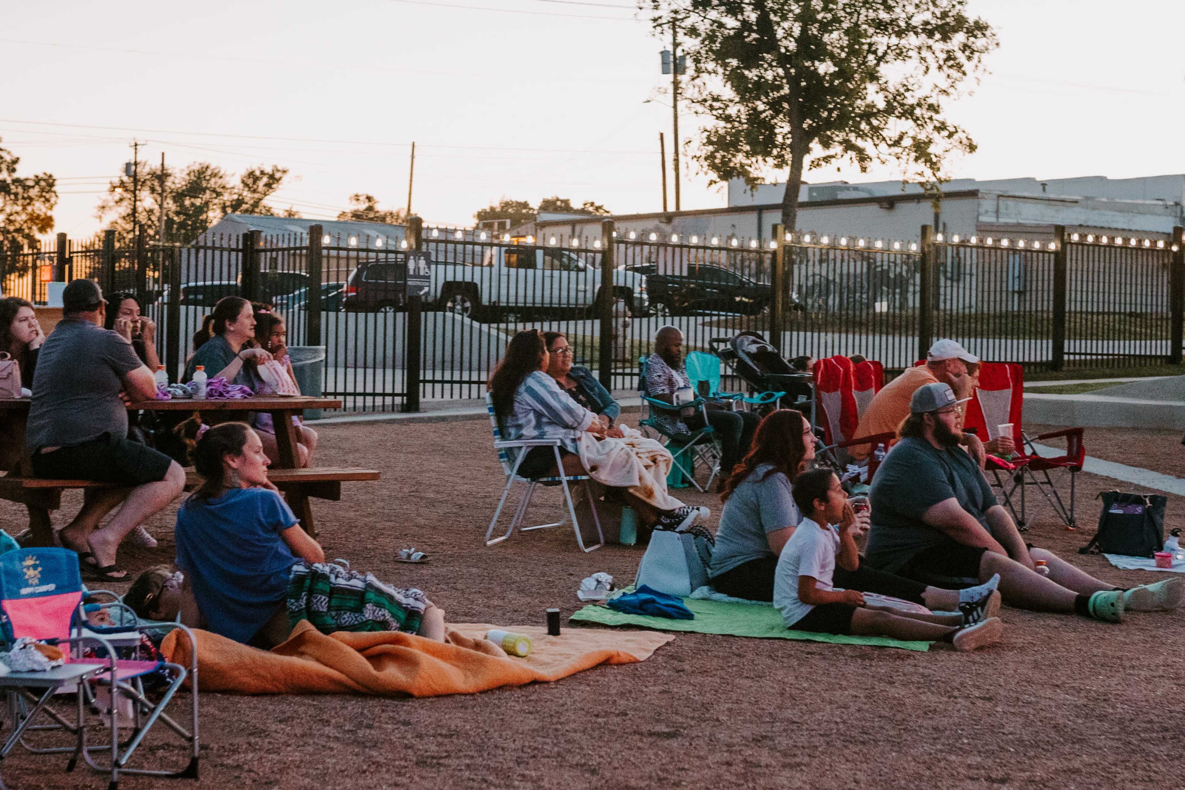 A group of people sit on blankets and chairs in an outdoor area, watching a movie projected on a screen as evening approaches.