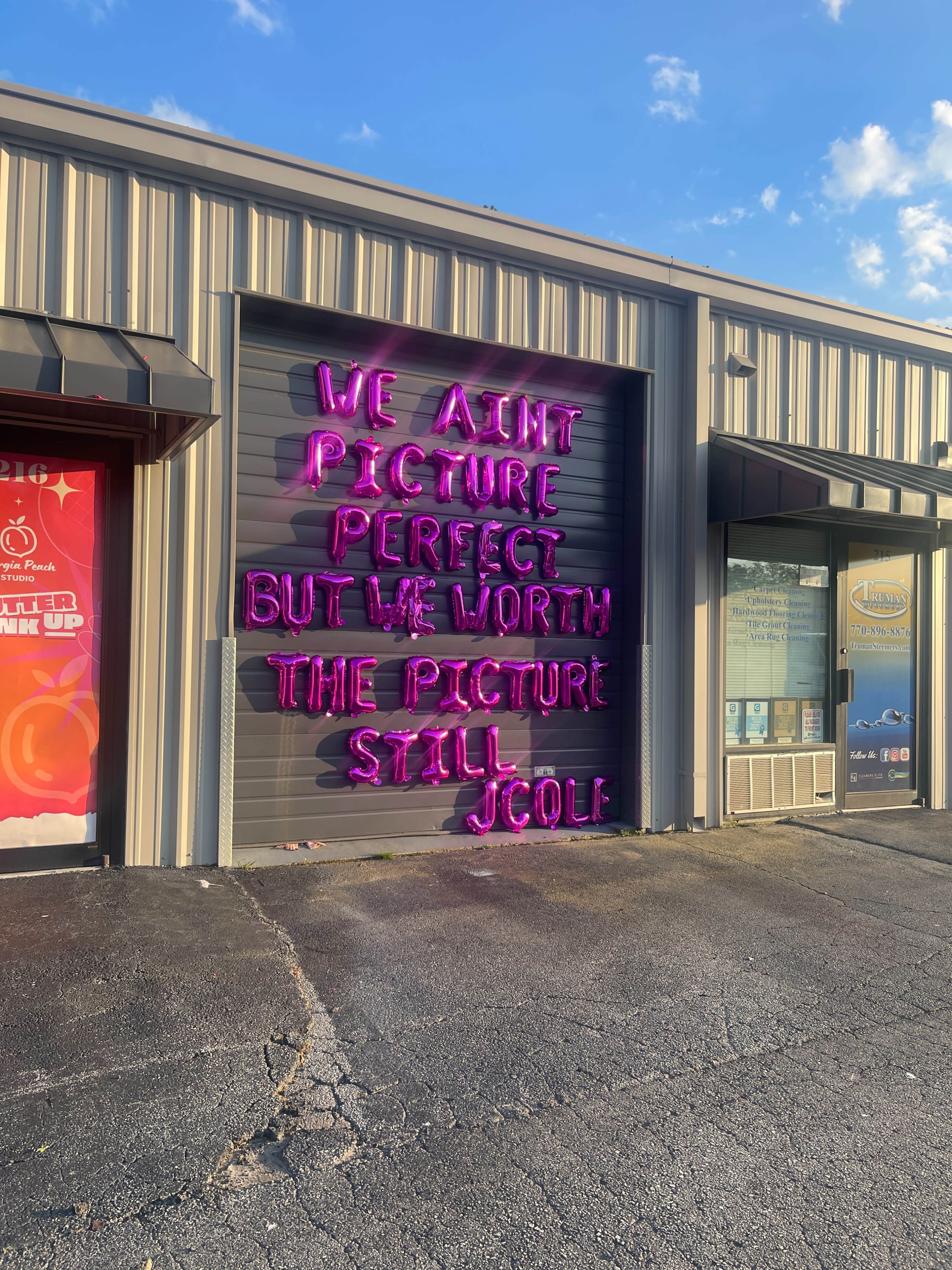The image shows a closed garage door with neon pink lettering that reads, "WE AINT PICTURE PERFECT BUT WE WORTH THE PICTURE STILL JCOLF."