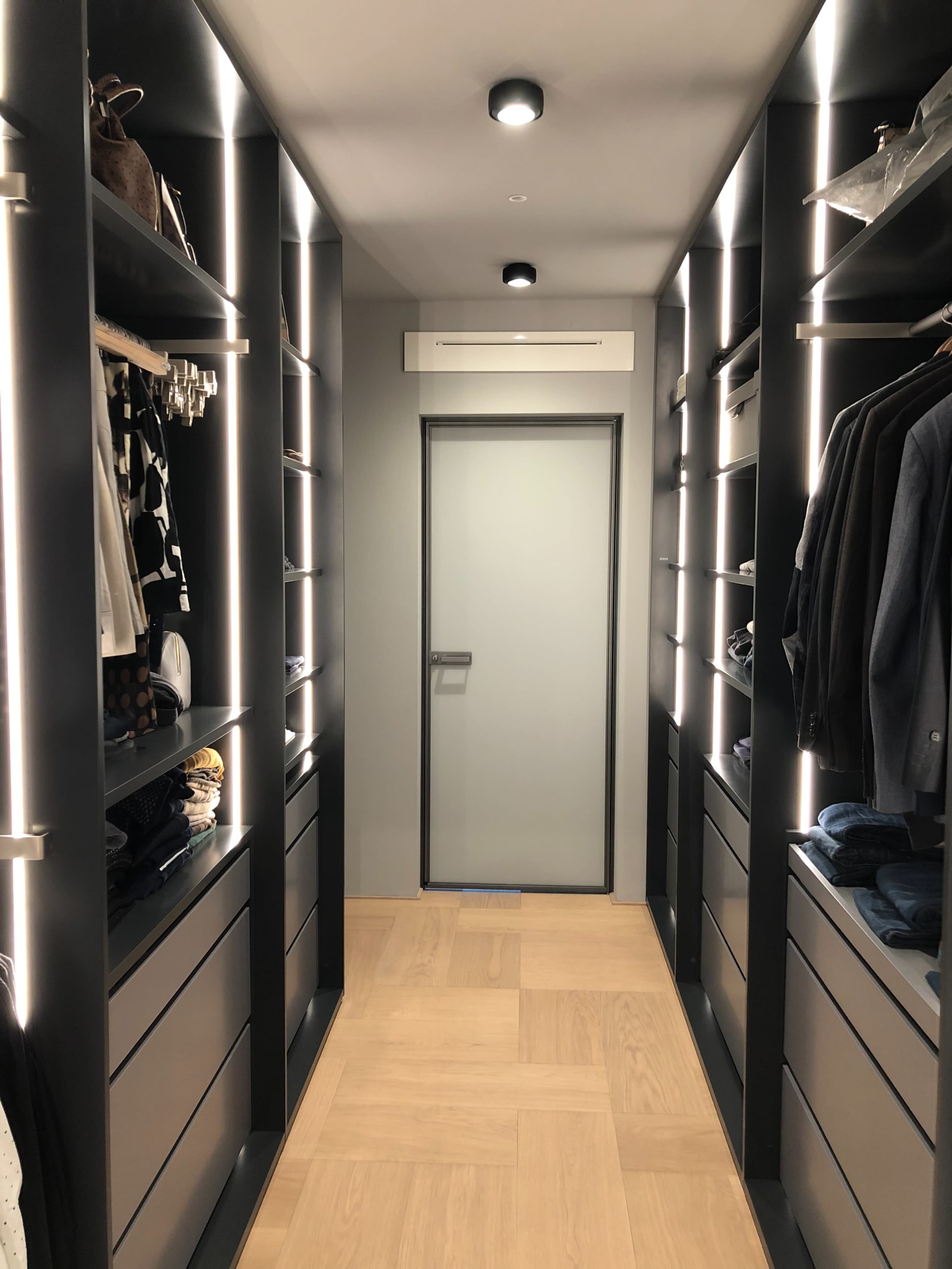 A narrow hallway with built-in shelves on both sides, illuminated by LED lights, leading to a gray door at the end.