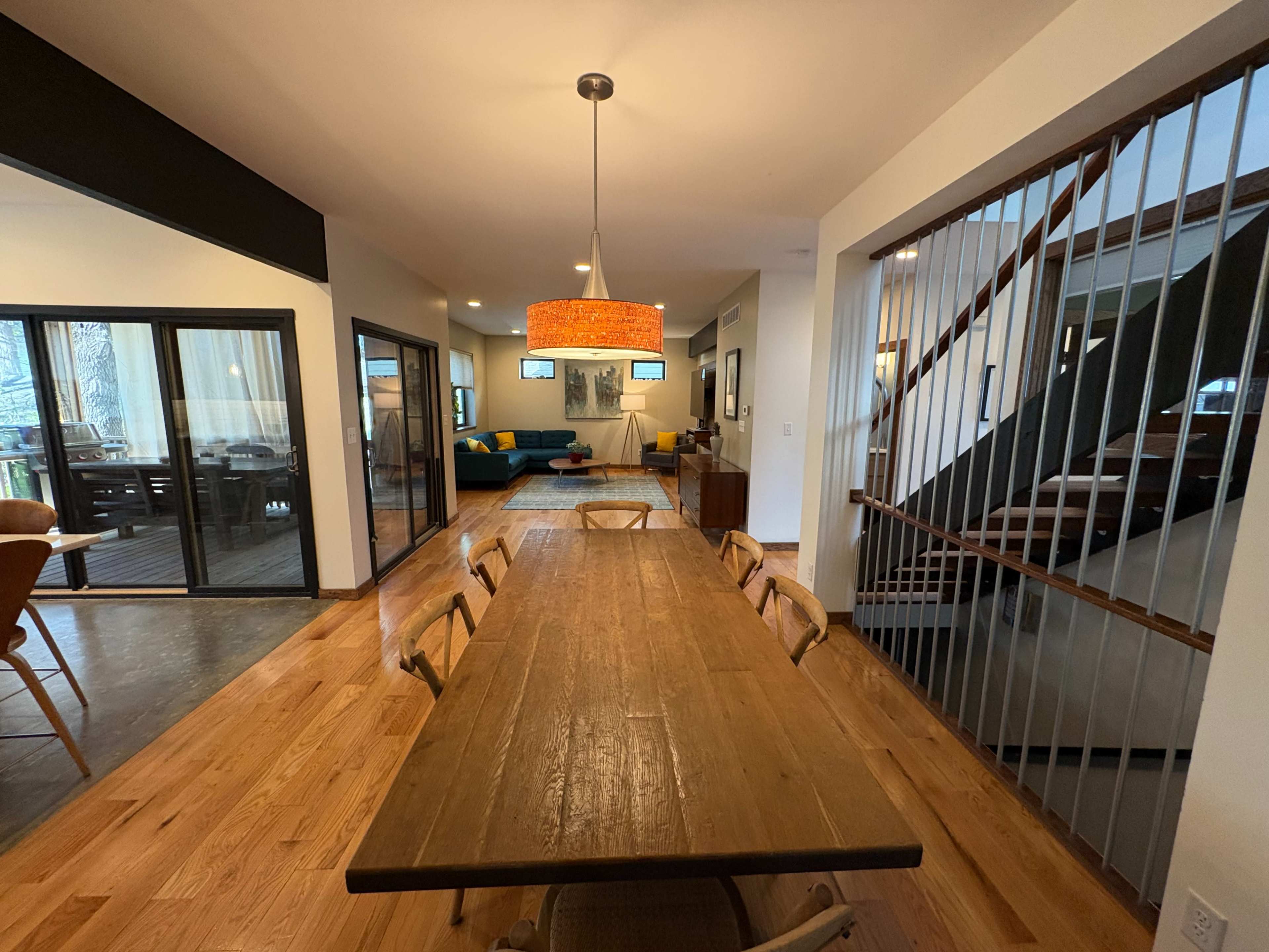 The image shows a dining area with a long wooden table, a pendant light fixture, and an open view toward a living space featuring a blue couch.