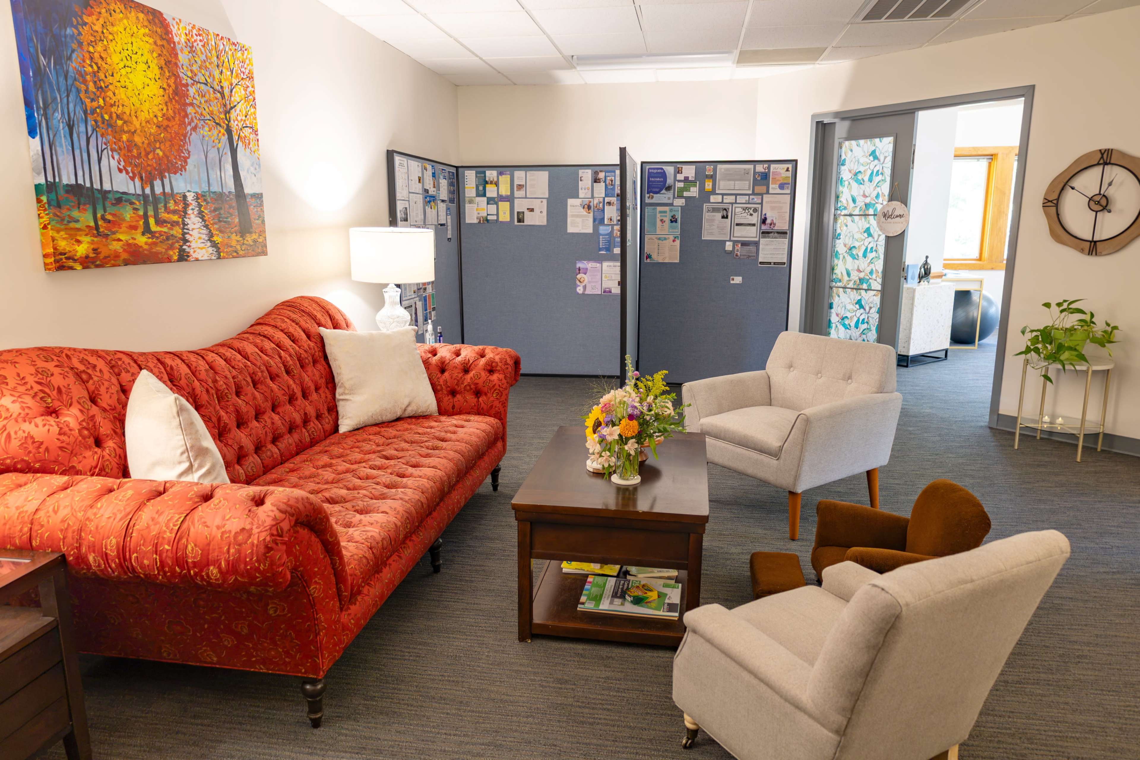 The image shows a furnished room with a red upholstered sofa, two armchairs, a coffee table with a flower arrangement, and informational boards in the background.