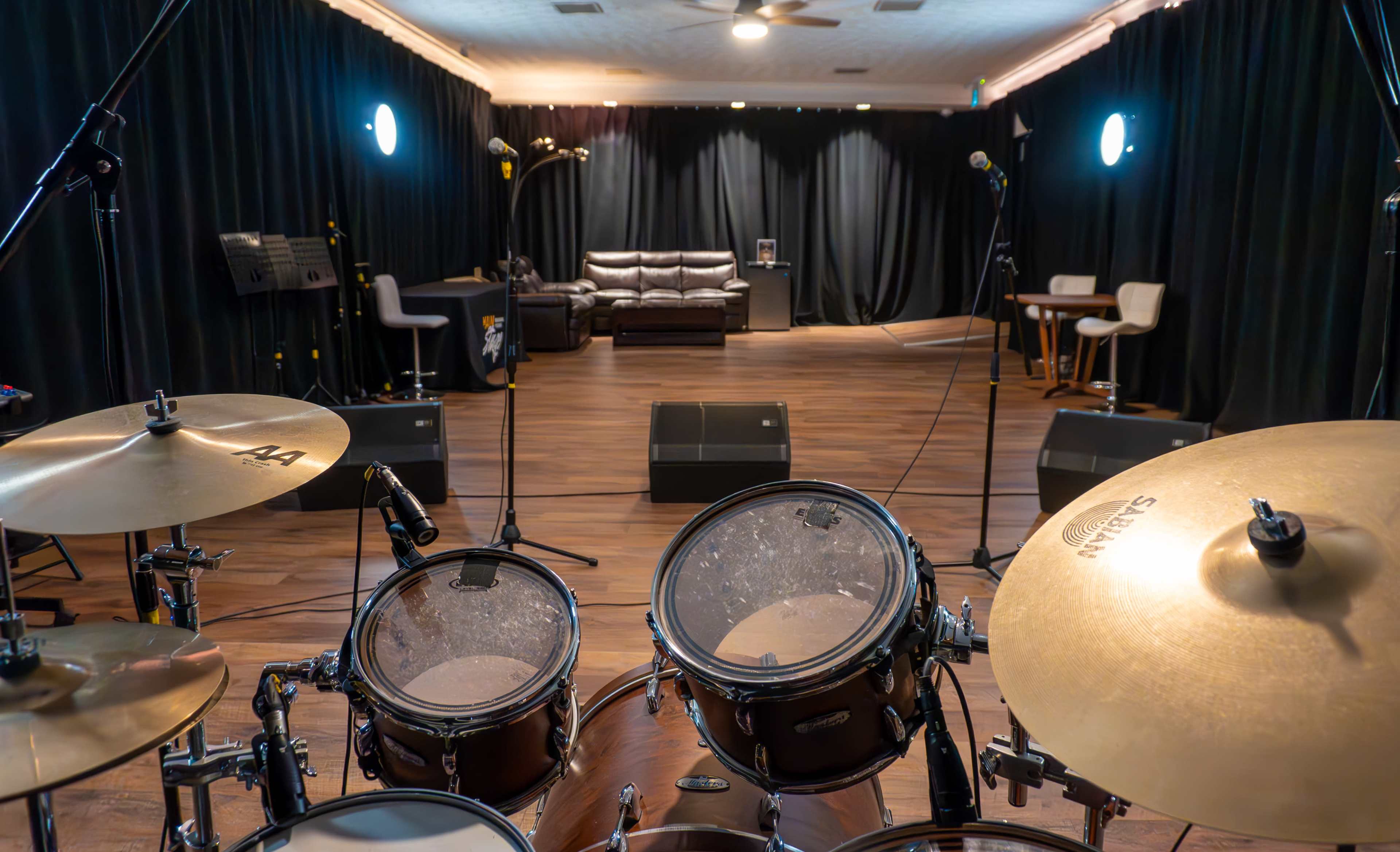 A drum set is positioned in front of a stage area with a sofa, chairs, and microphones, all framed by dark curtains.