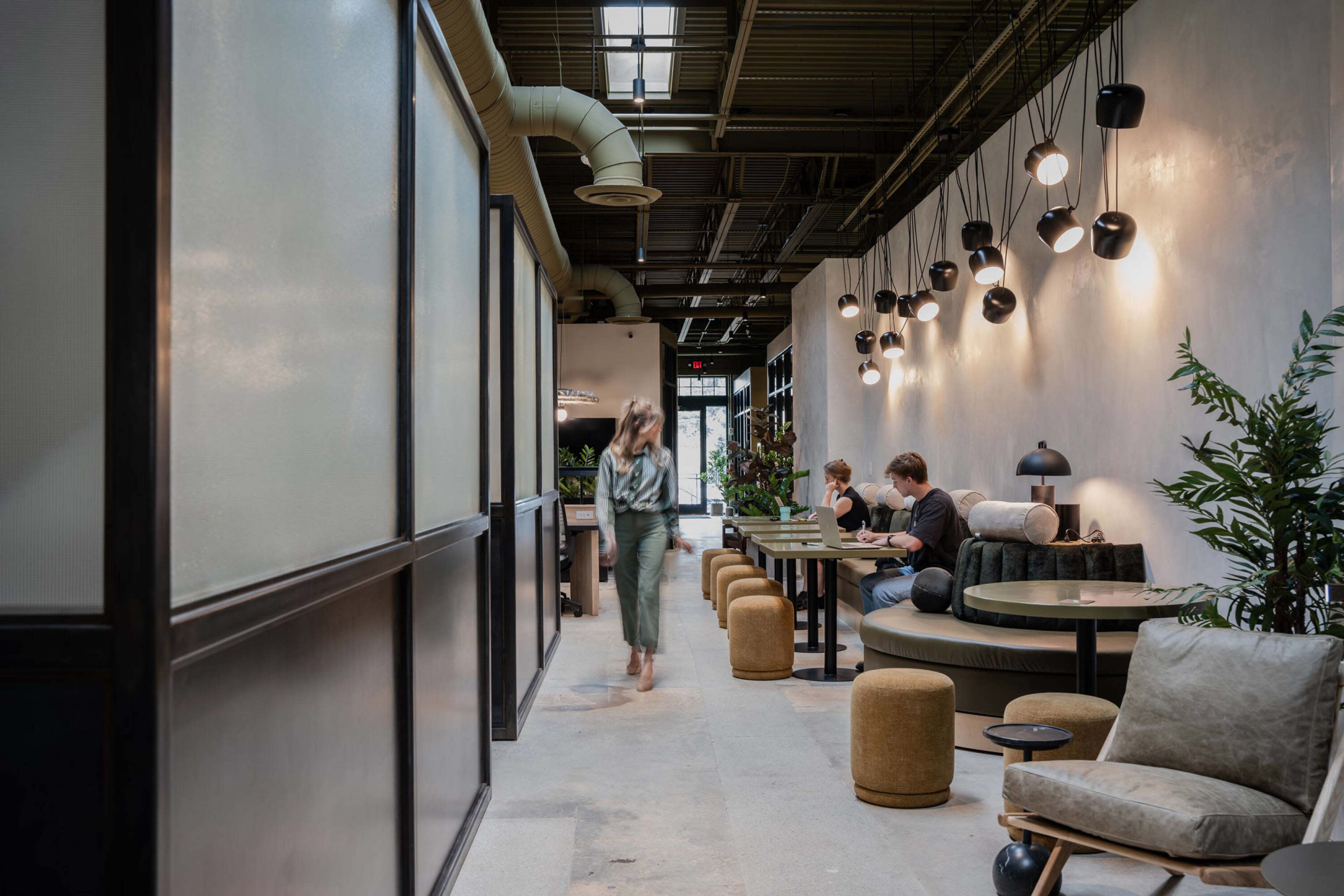 A modern indoor space features a narrow hallway with seating areas, plants, and pendant lighting, where individuals are engaged in conversation or work.