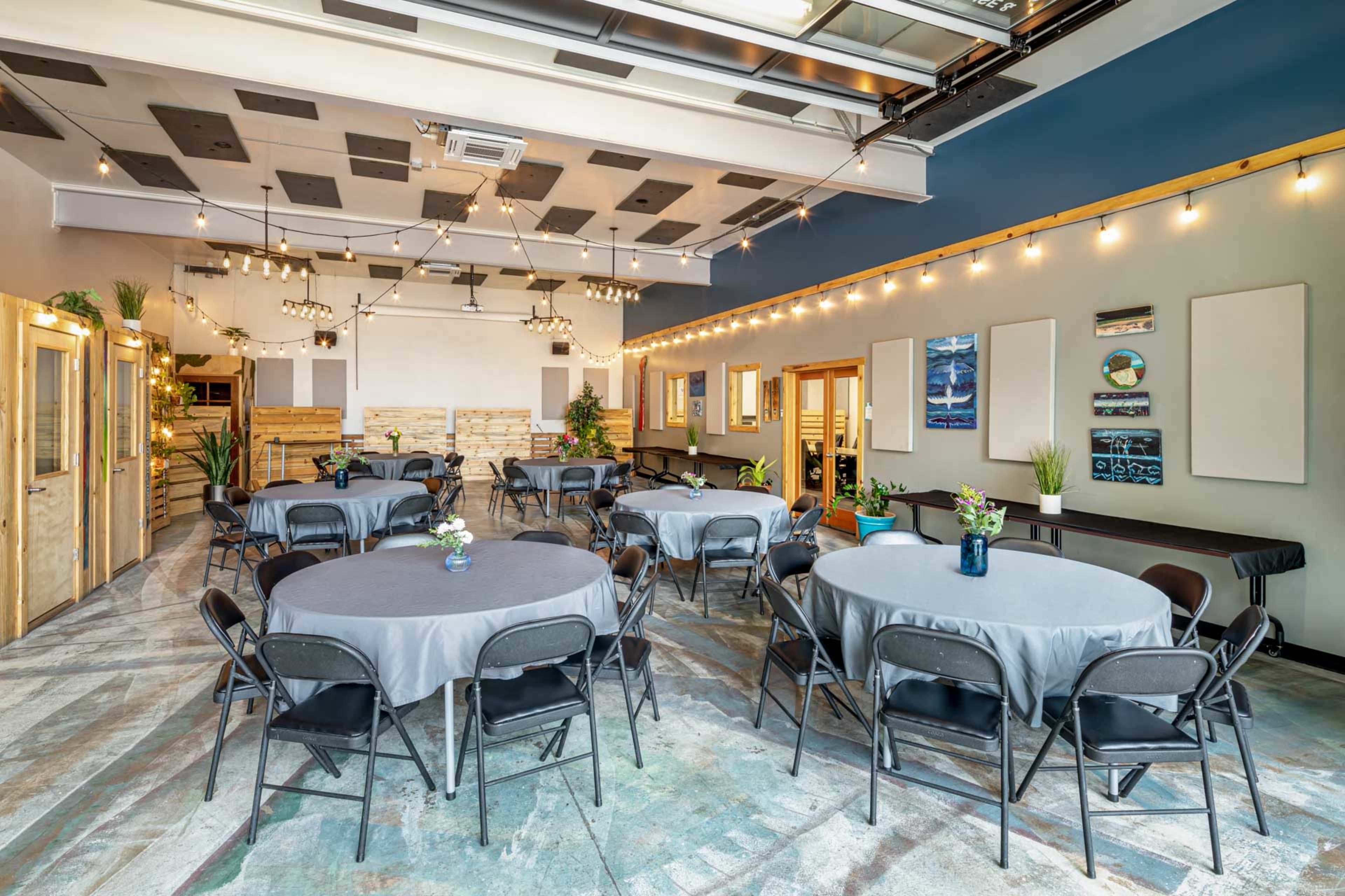 The image shows a spacious event room with round tables covered in gray tablecloths, string lights overhead, and wooden elements in the decor.