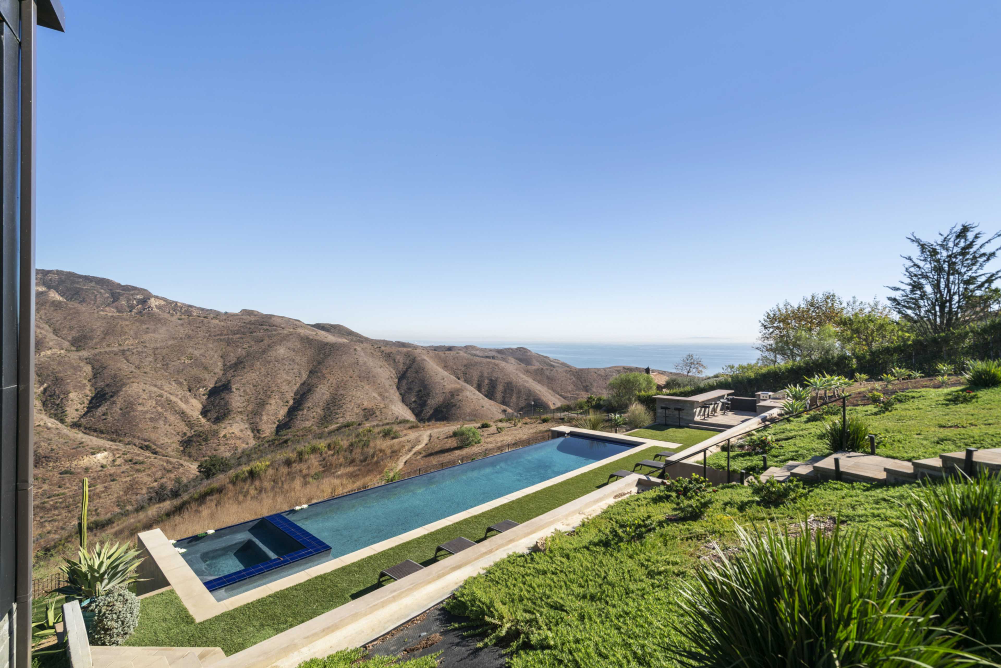 A modern house overlooking a long rectangular swimming pool and rolling hills leading to the ocean in the distance.