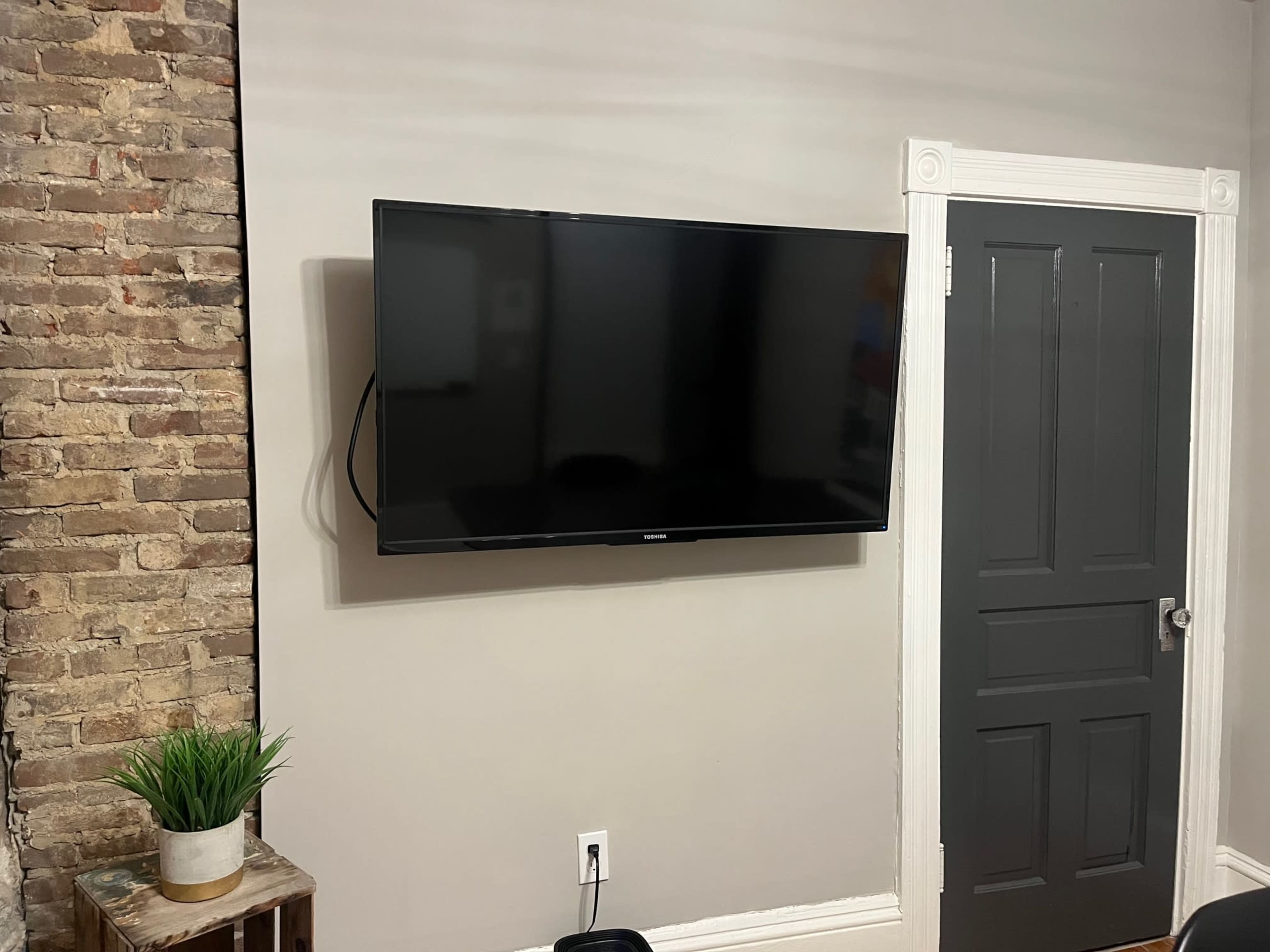 A flat-screen television is mounted on a gray wall next to a brick section of the wall and a closed gray door.