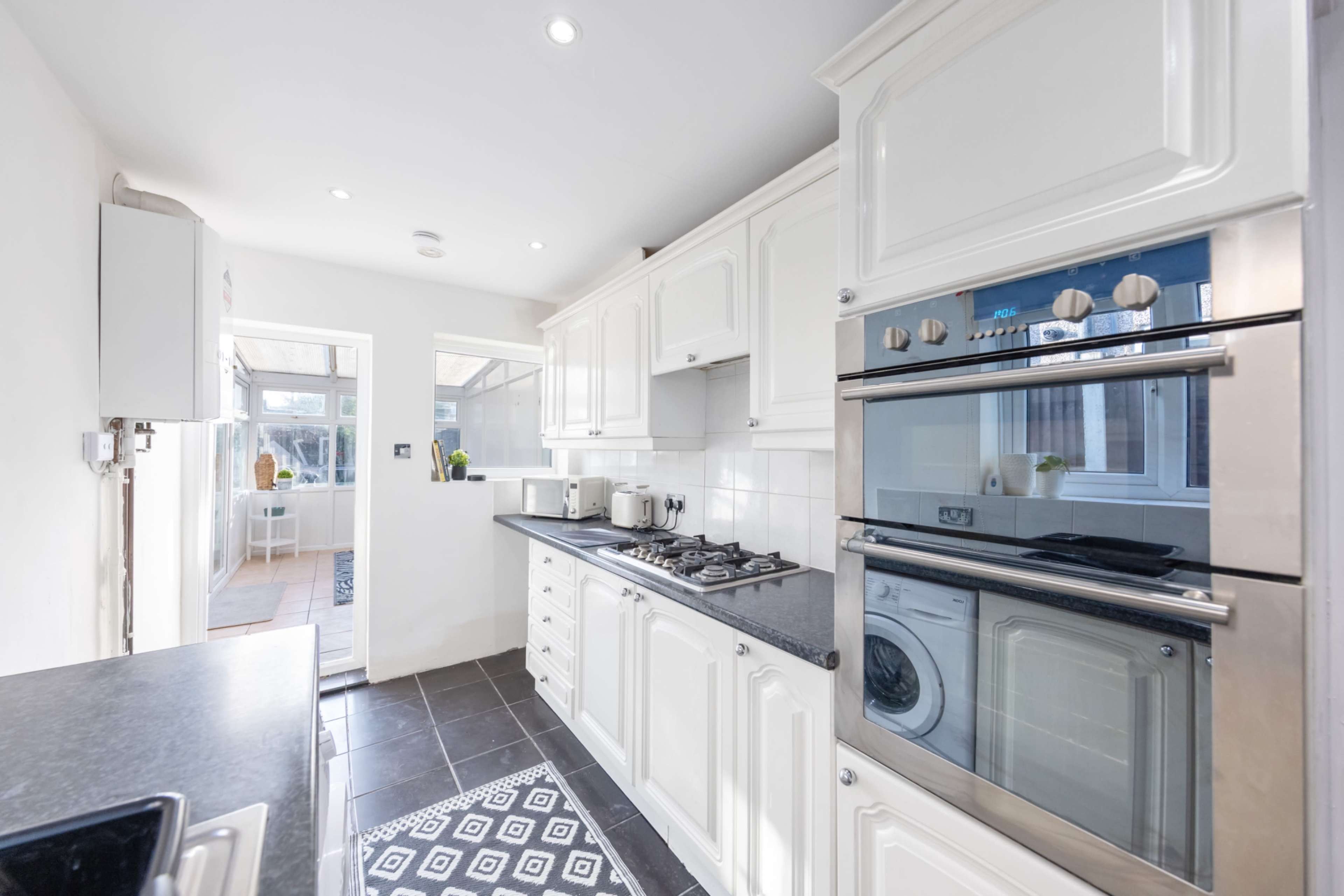 The image shows a modern kitchen with white cabinetry, stainless steel appliances, a gas stove, and black countertops, featuring tiled floors and a window leading to an adjoining area.