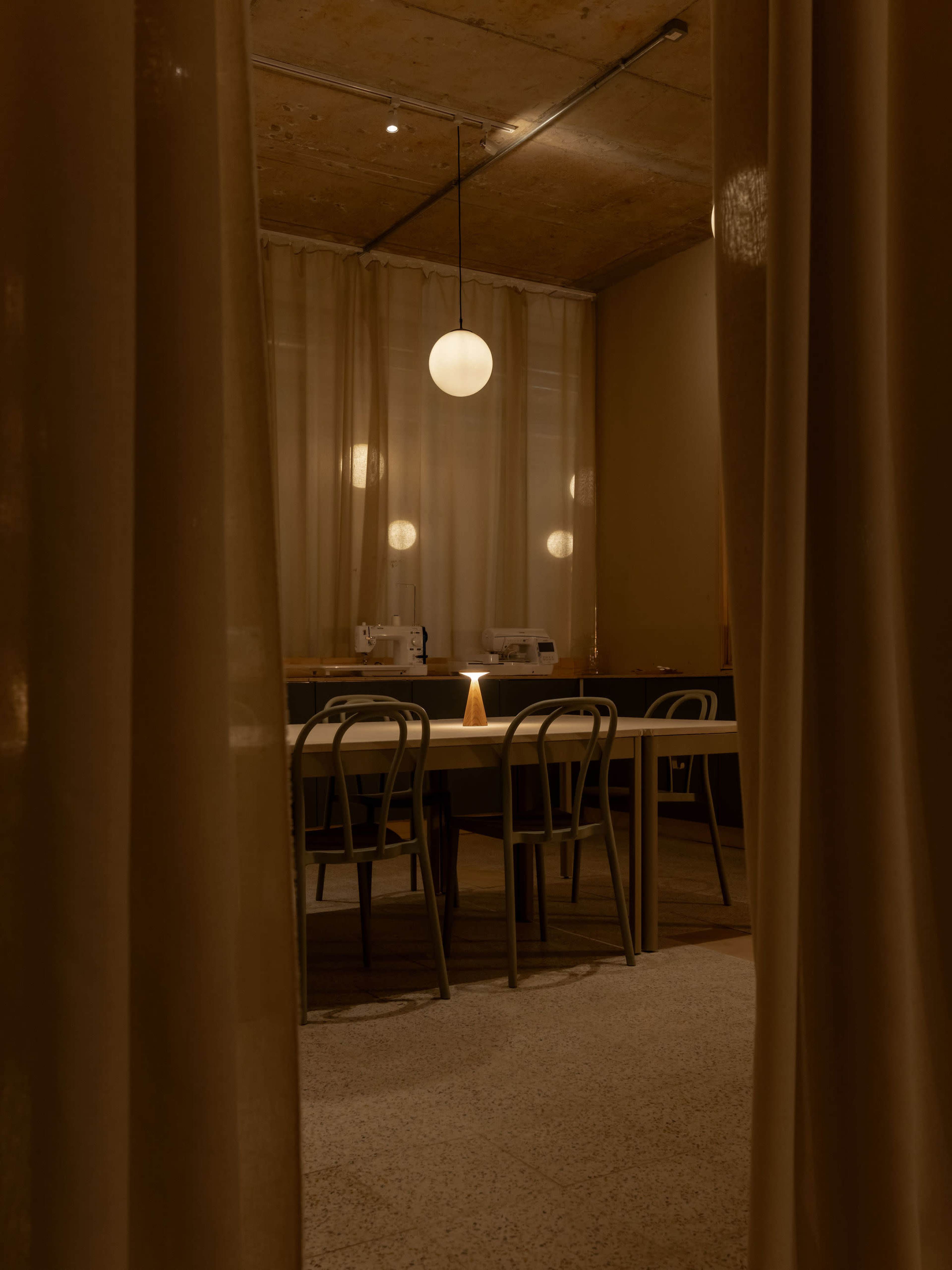 A softly lit room with a large table surrounded by metal chairs and sewing machines visible in the background, framed by flowing curtains.