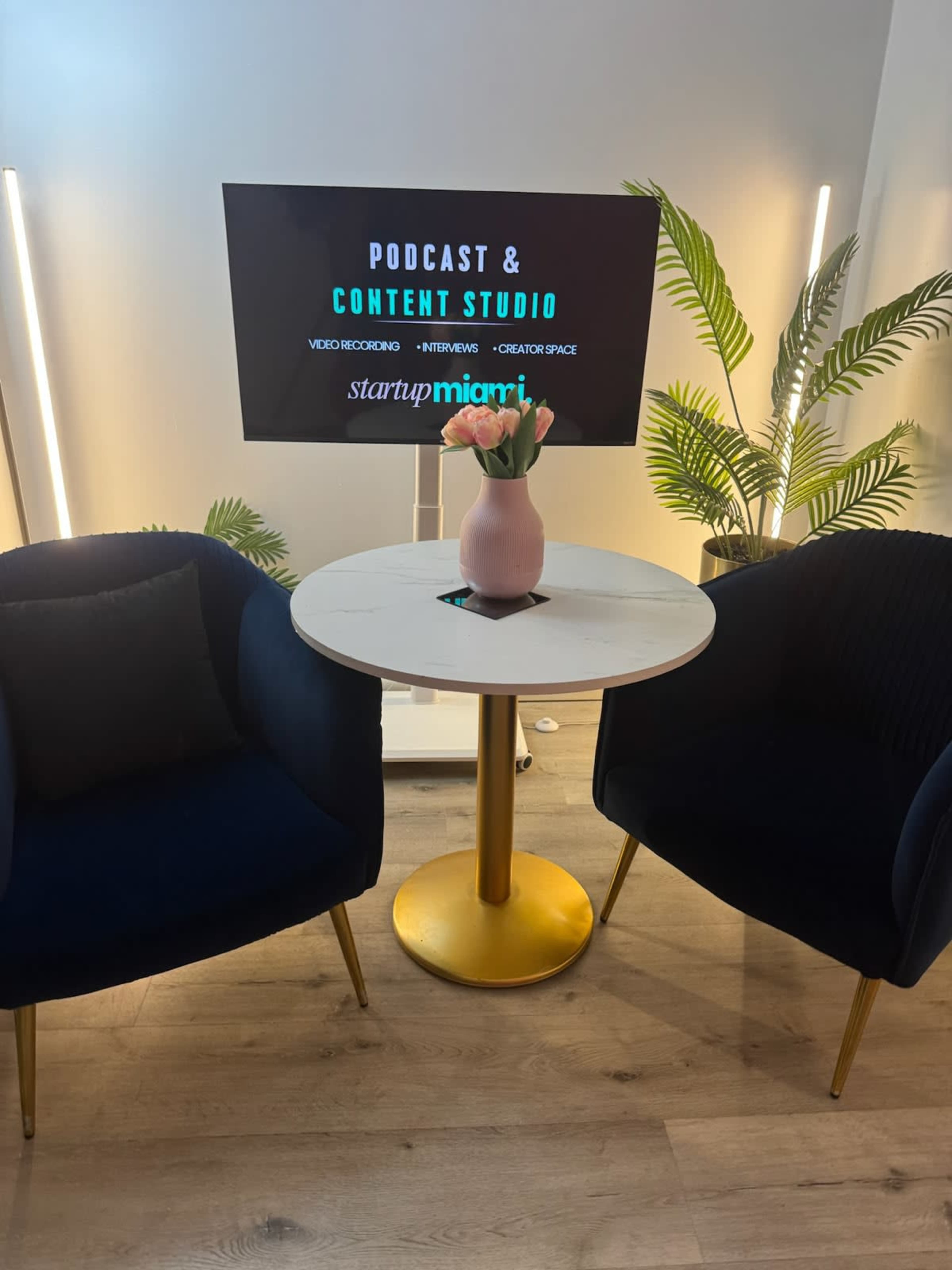 The image shows a small table with a vase of flowers between two blue chairs, with a screen in the background displaying the text "PODCAST & CONTENT STUDIO" and "startupmiami."