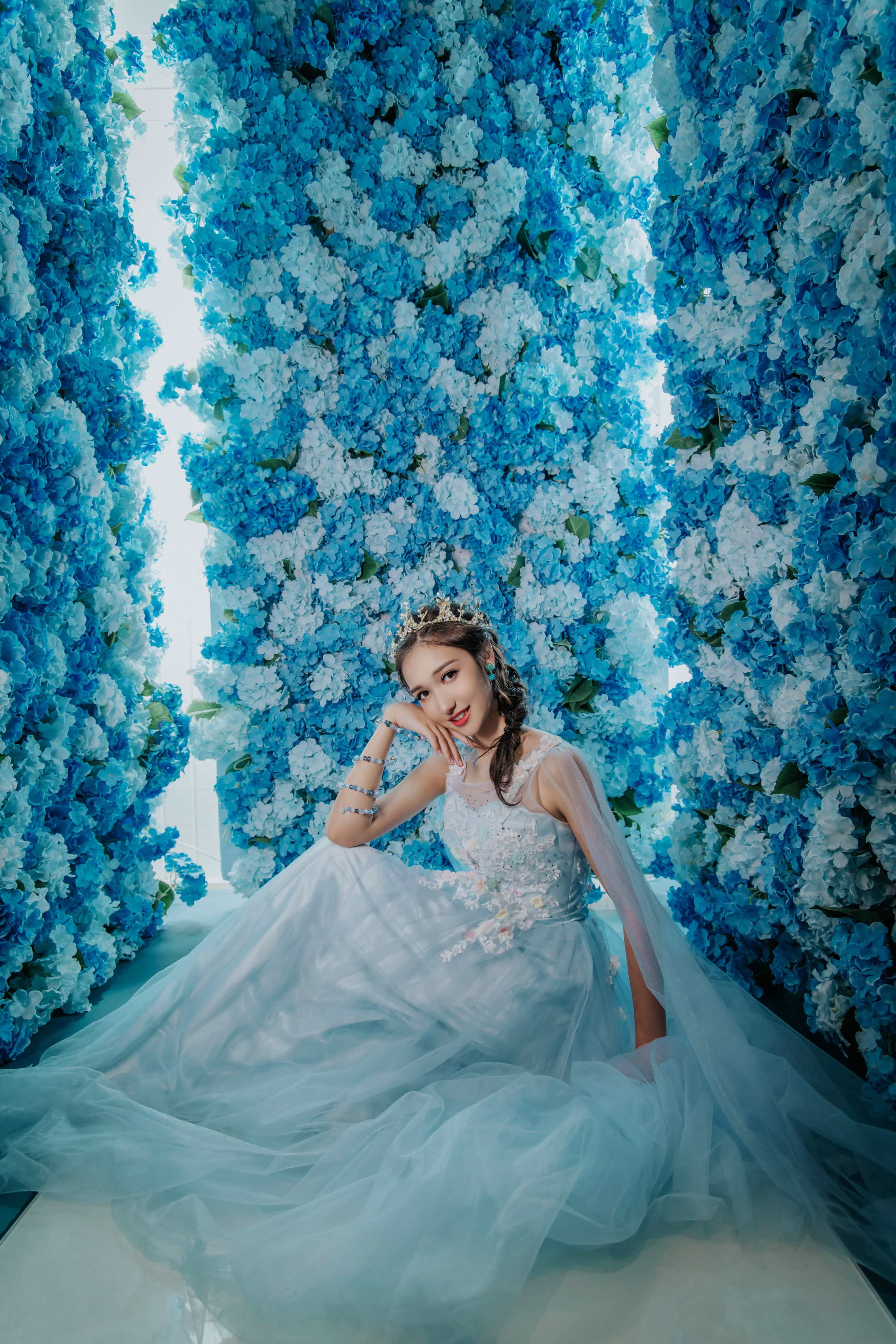A girl in a flowing dress poses among tall arrangements of blue and white flowers.