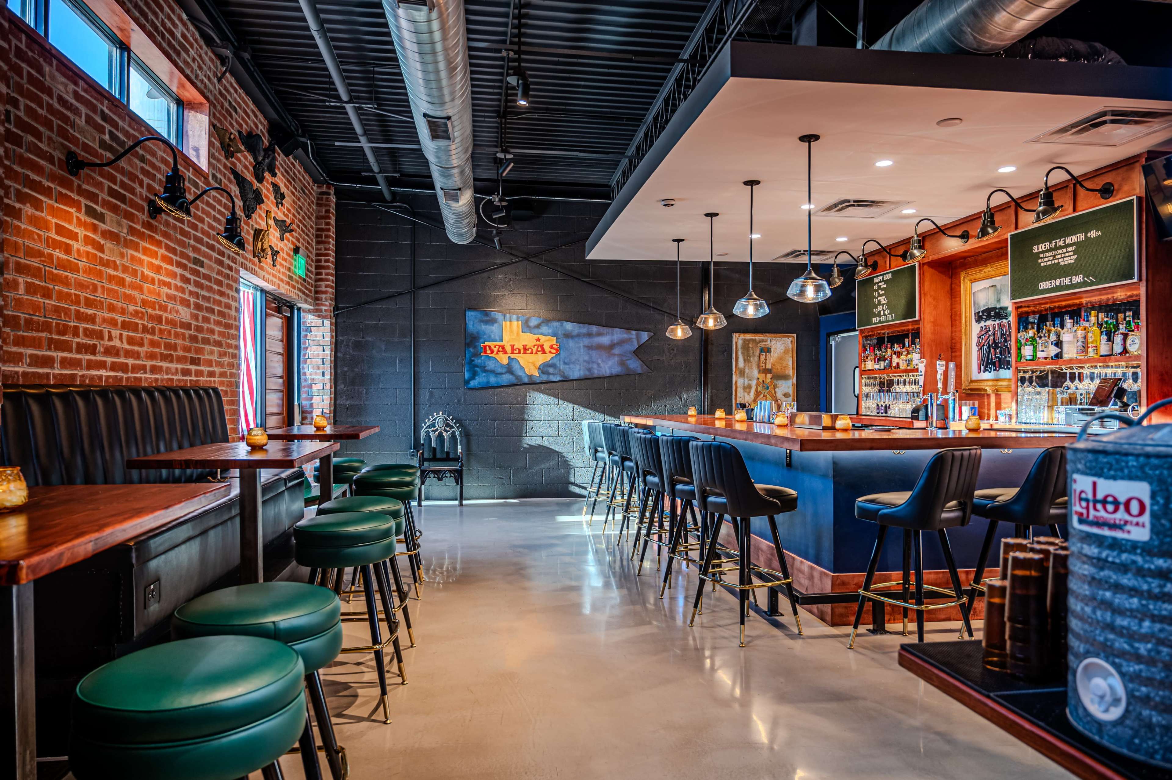 The interior of a modern bar features dark green and wooden accents, a long bar counter with stools, and a mix of high-top and low seating arranged against exposed brick walls.