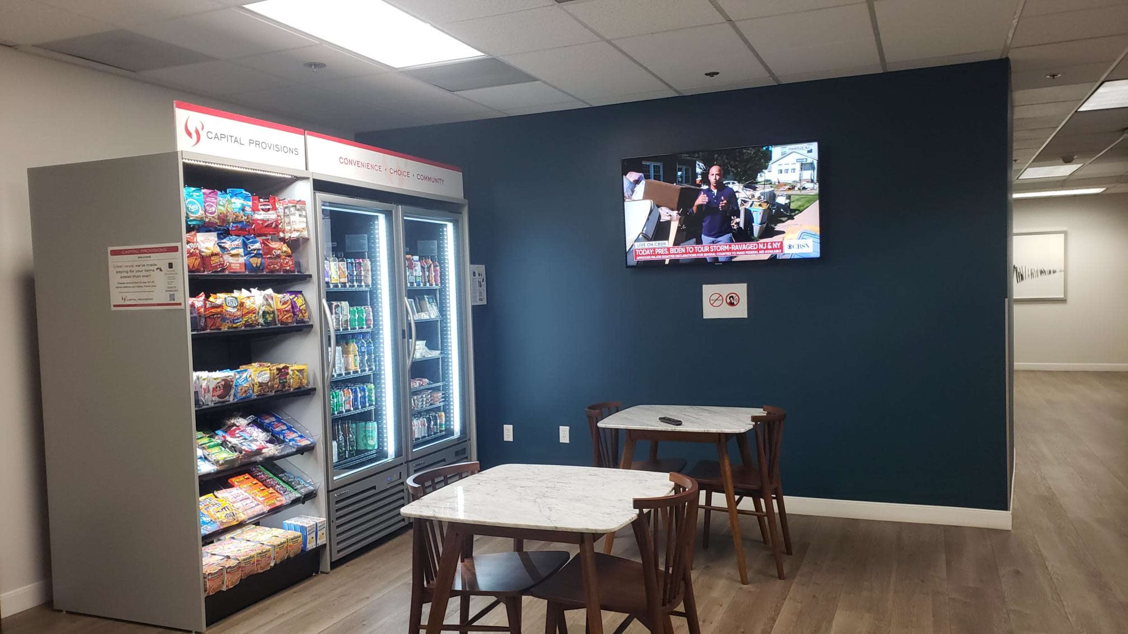 A break area with a vending machine filled with snacks and beverages, two wooden tables, and a television mounted on the wall displaying news.