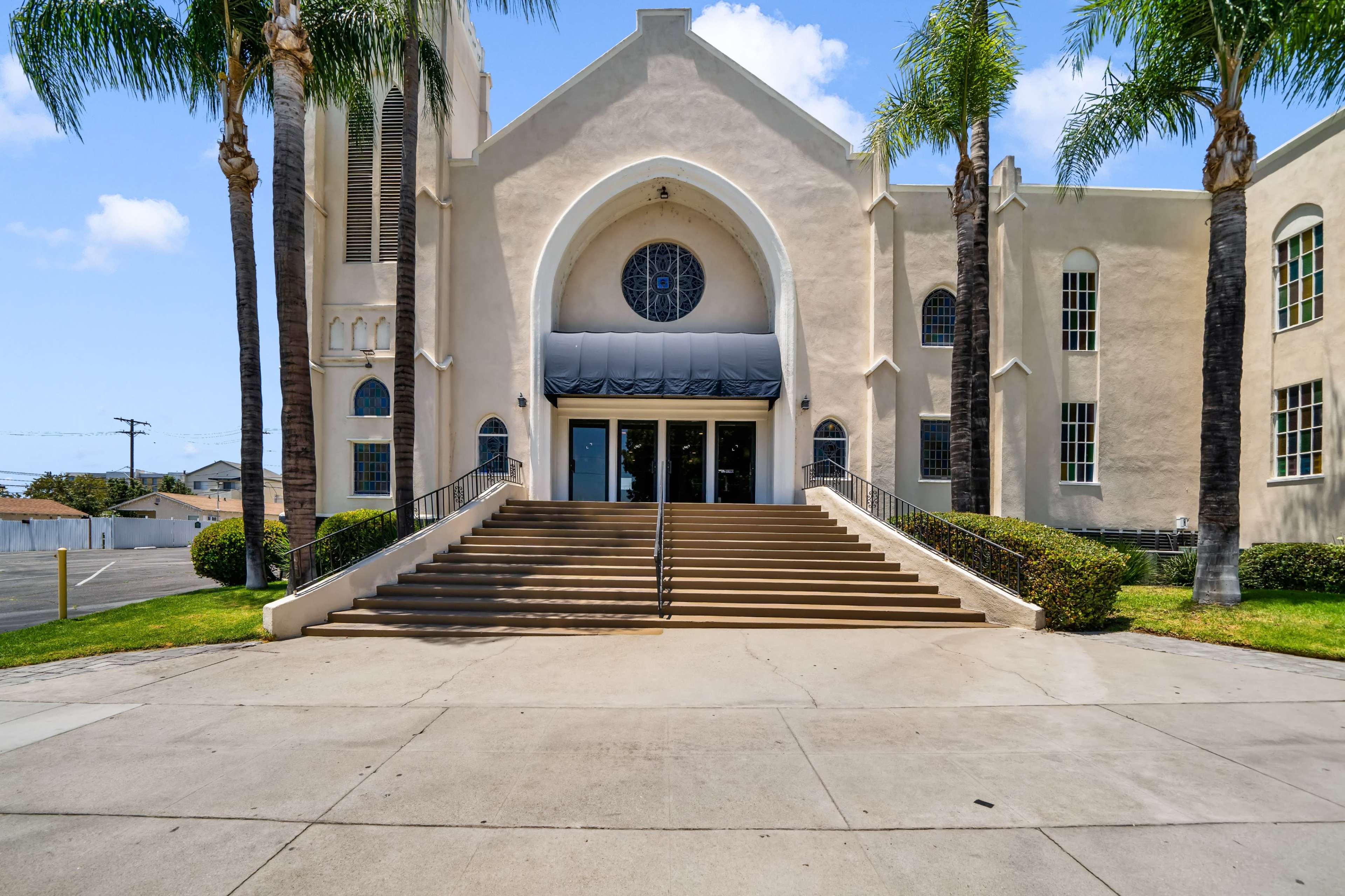 The image shows the front entrance of a building with a large staircase flanked by palm trees, featuring a circular window above the main doors.