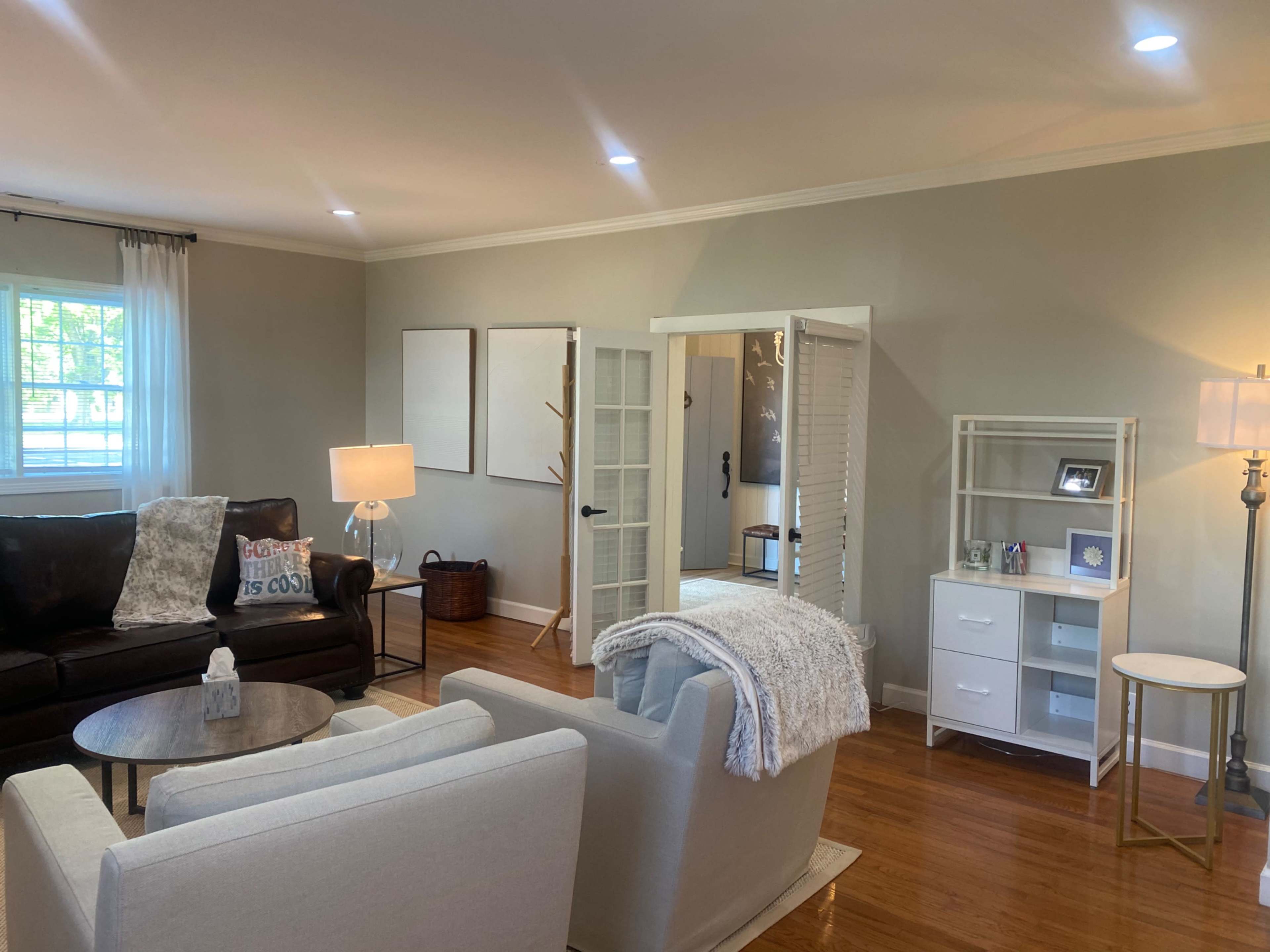 The room features a cozy living space with a dark sofa, light-colored armchairs, a white bookshelf, and sliding doors leading to another area.