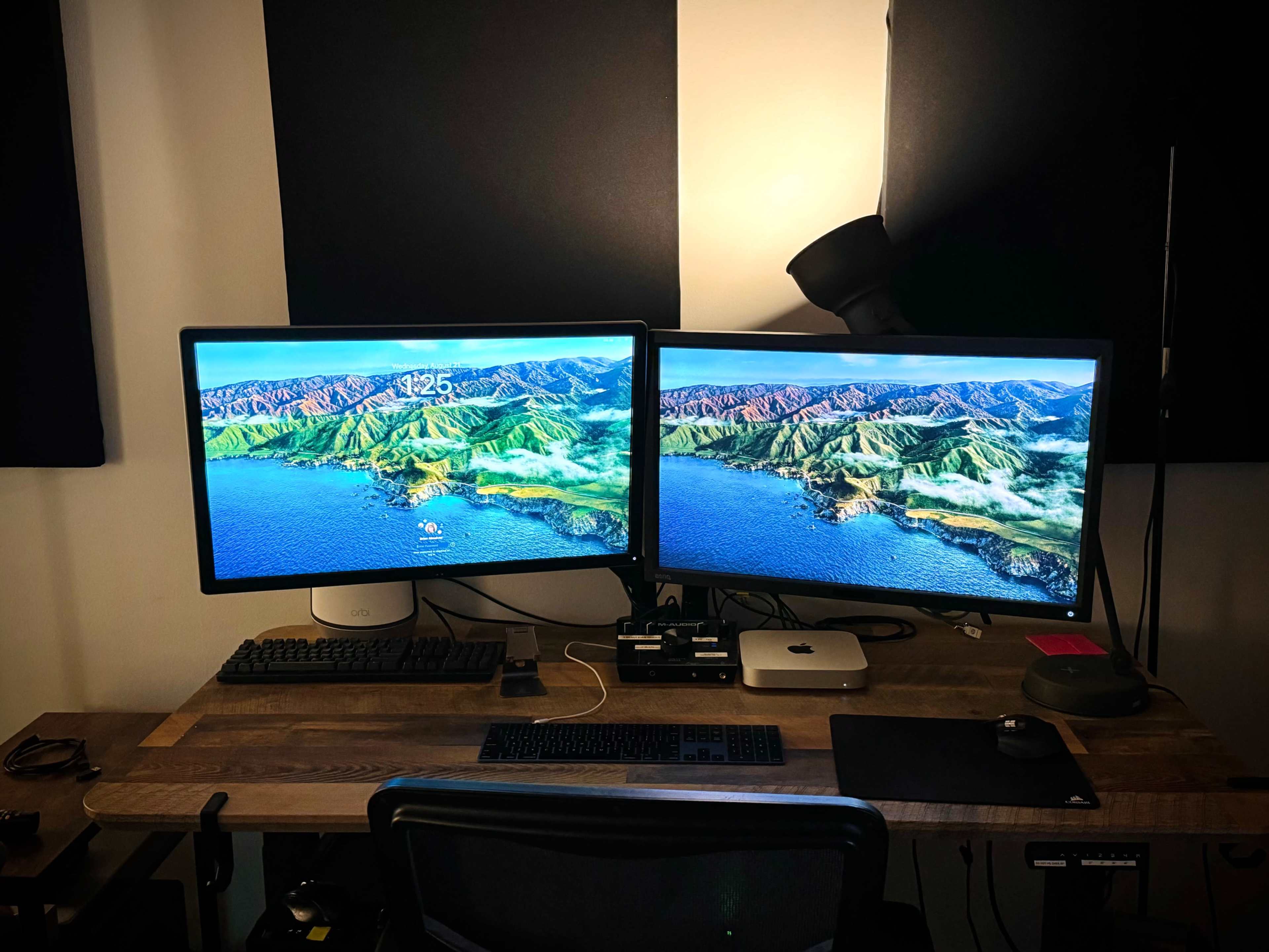 A workspace features two monitors displaying a landscape, a computer tower, and a black chair on a wooden desk.