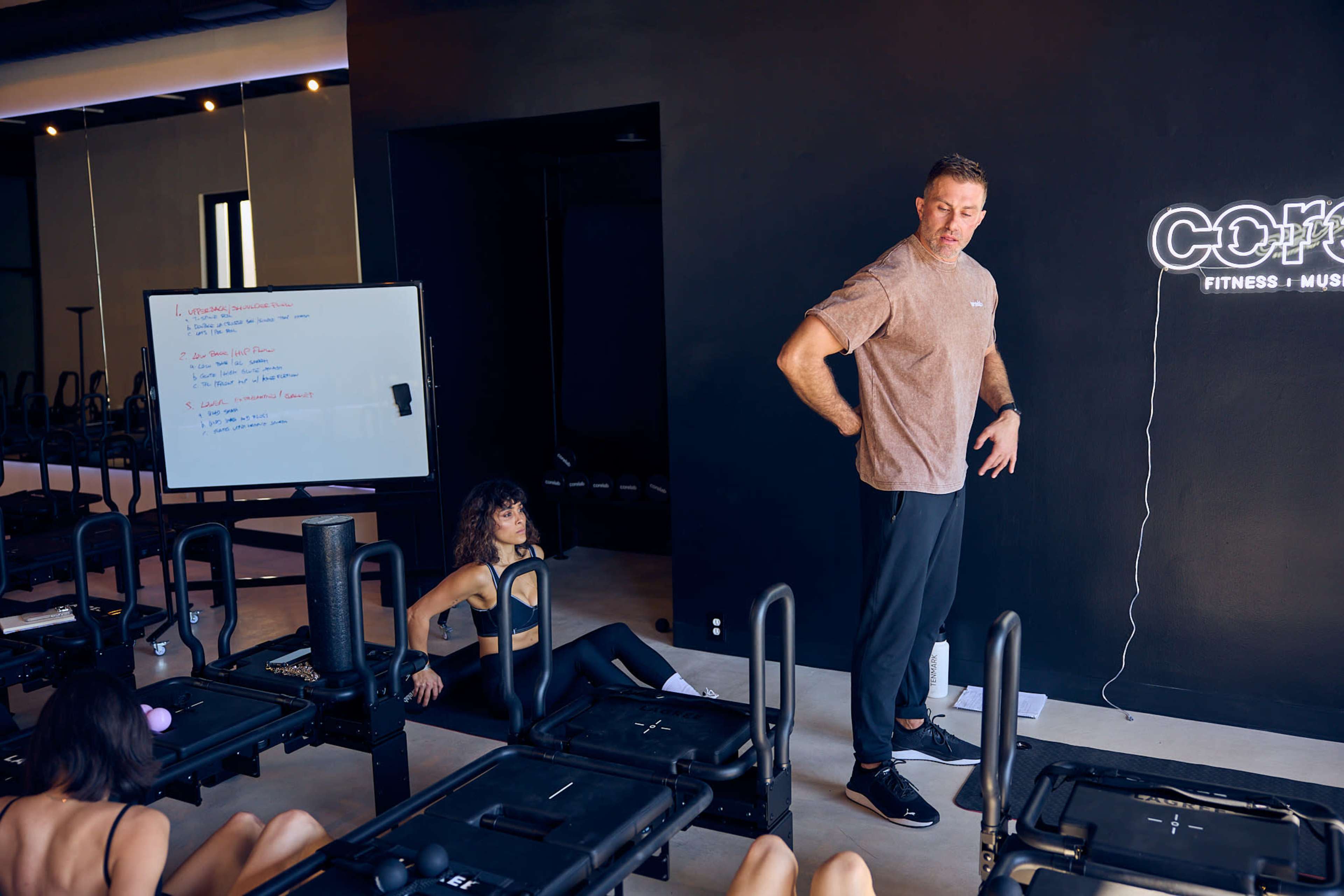 A fitness instructor stands in front of a group, addressing participants on exercise equipment in a modern gym studio.
