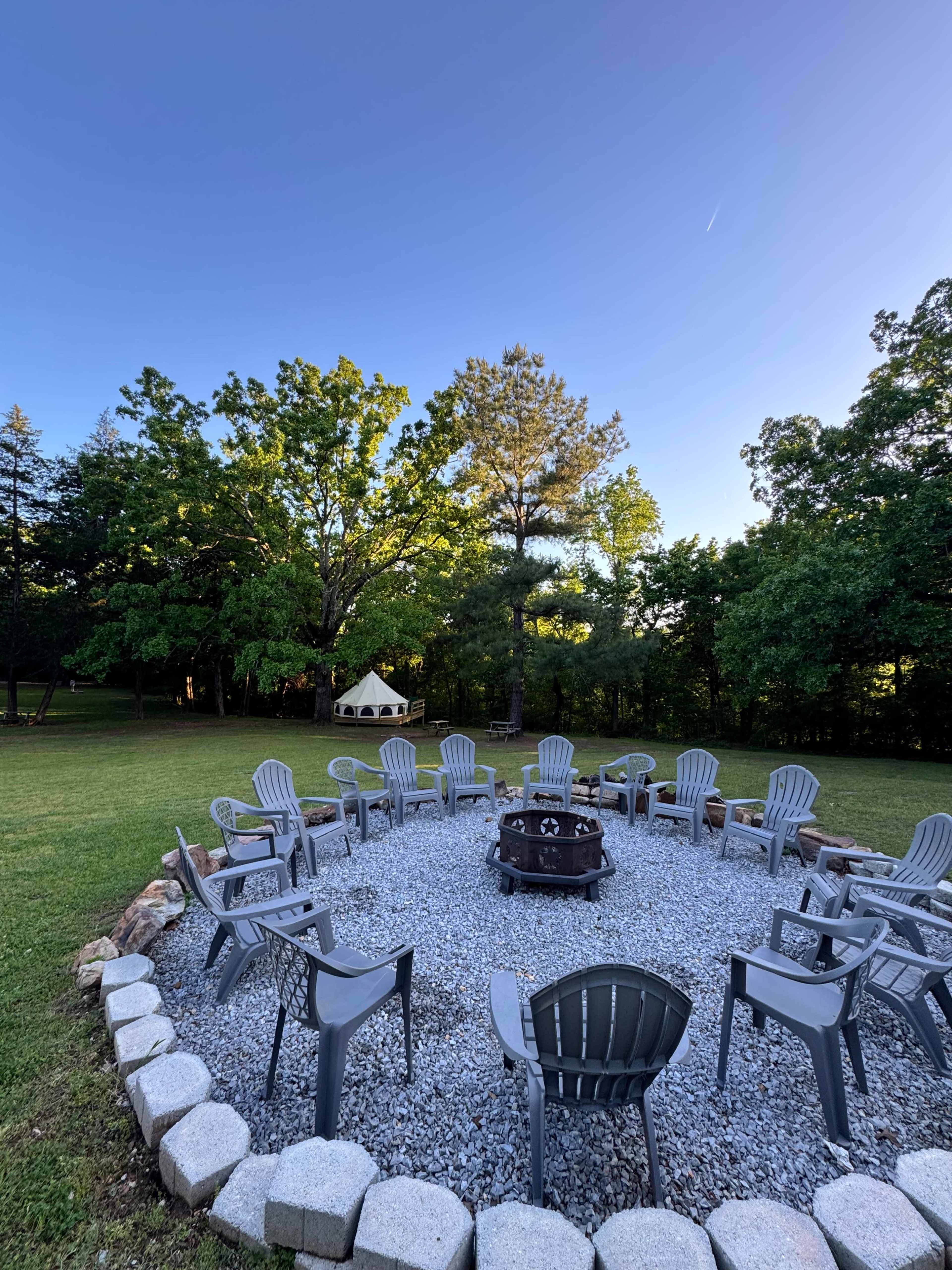 A circular arrangement of gray chairs surrounds a fire pit on a gravel surface in a grassy area with trees in the background.