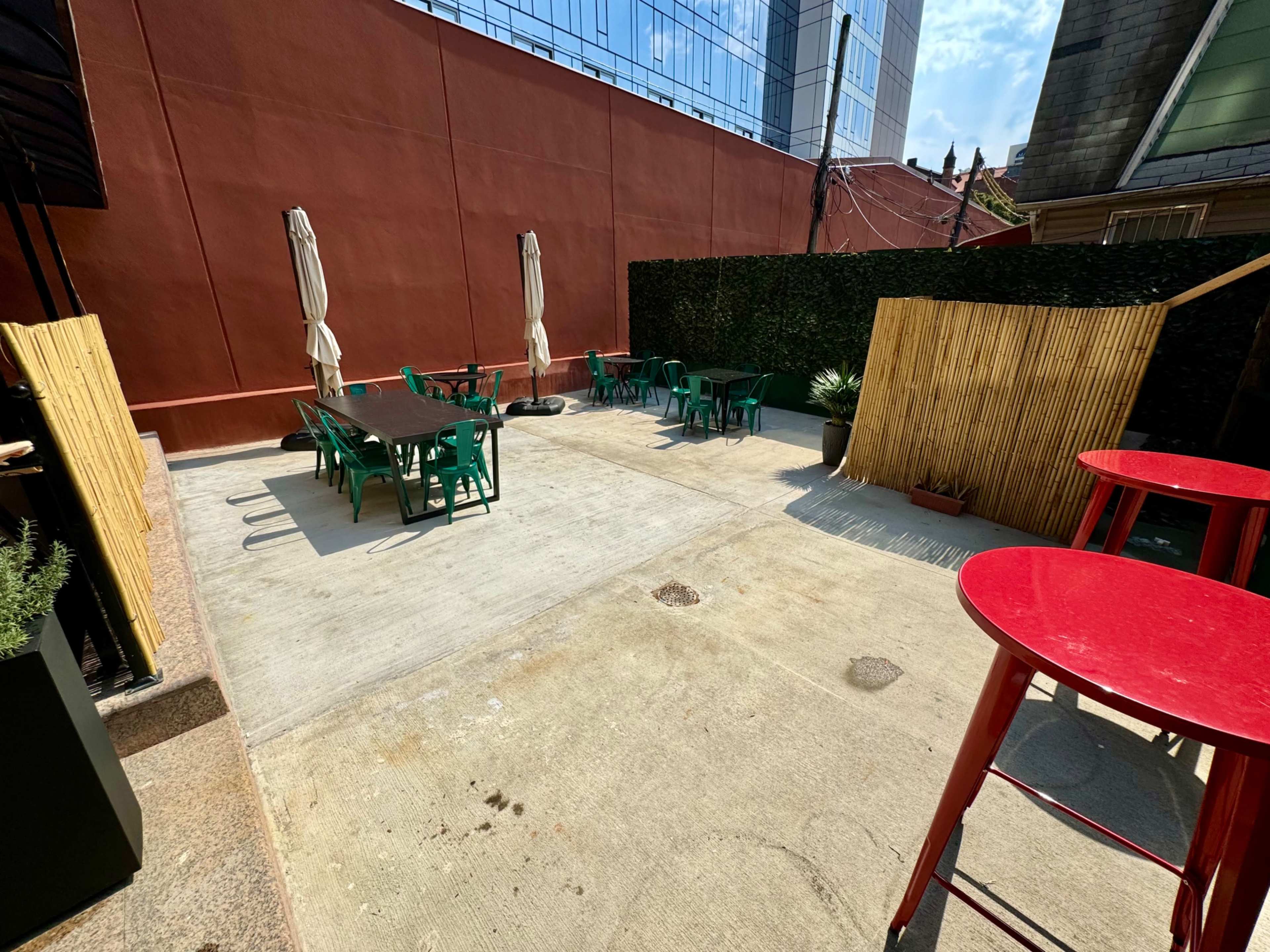 The image shows an outdoor patio area with green tables and chairs, surrounded by tall bamboo screens and large umbrellas.