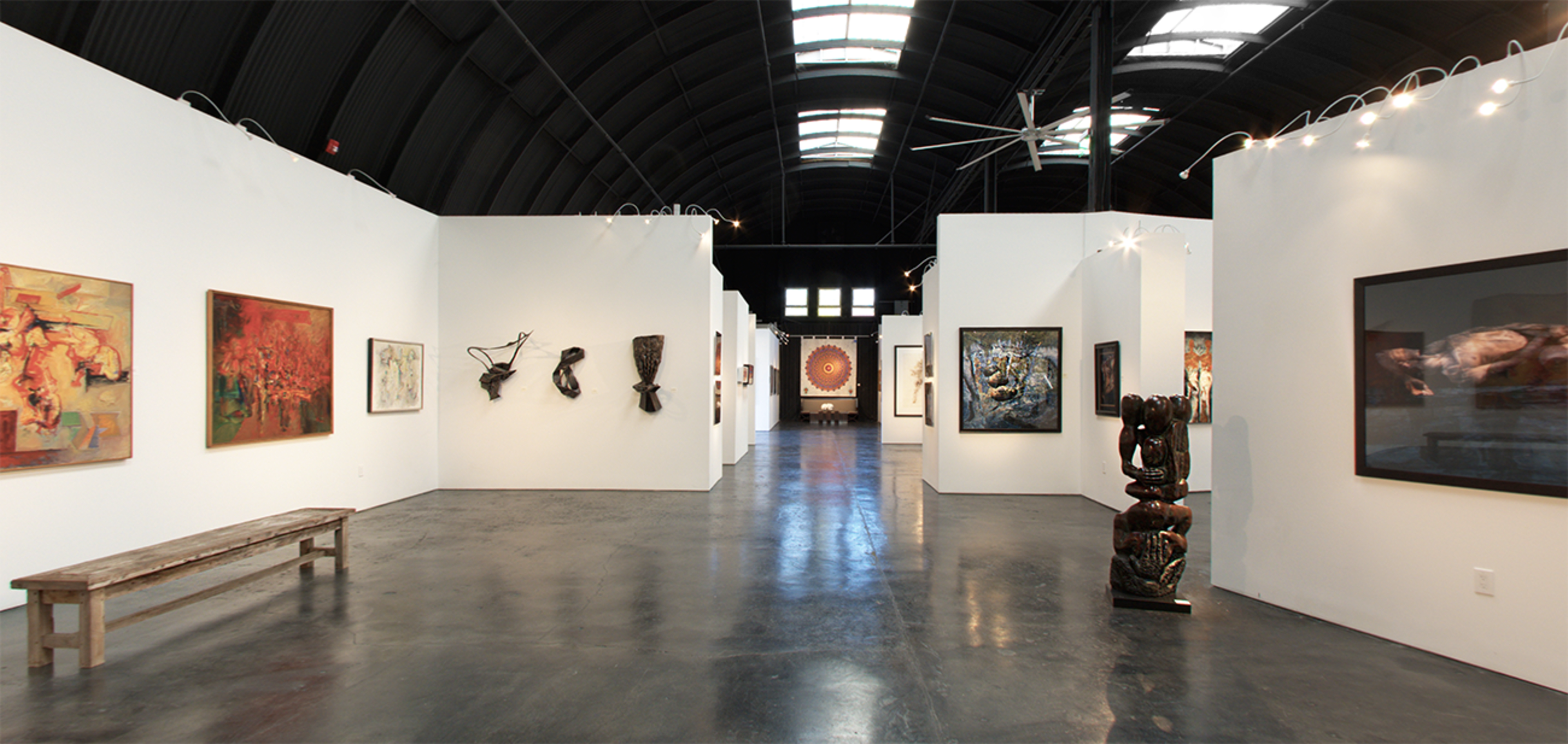 An interior view of an art gallery featuring various artworks displayed on white walls, with a polished floor and wooden bench visible in the foreground.