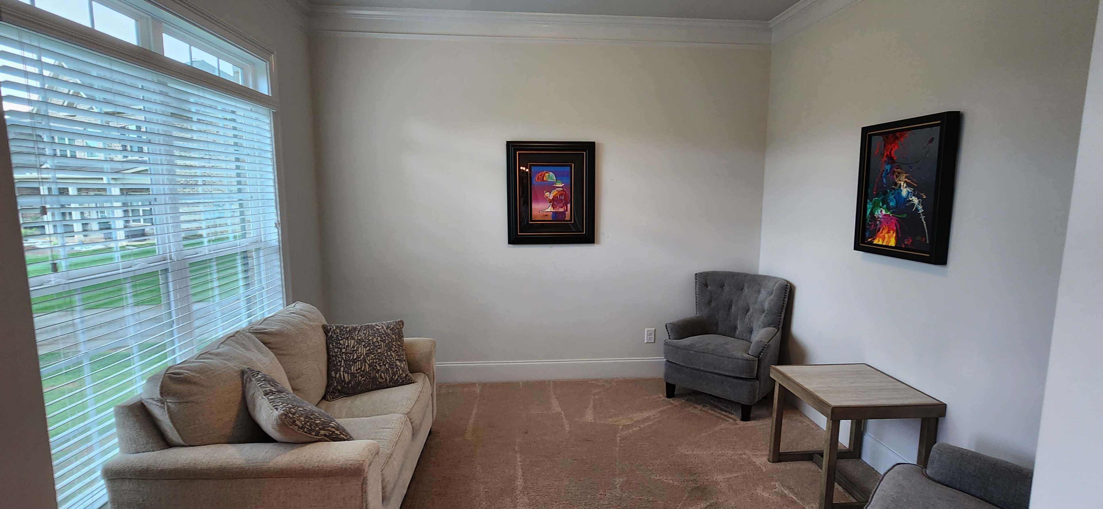 A small, well-lit room containing a beige sofa, a gray armchair, a side table, and two framed artworks on the wall.