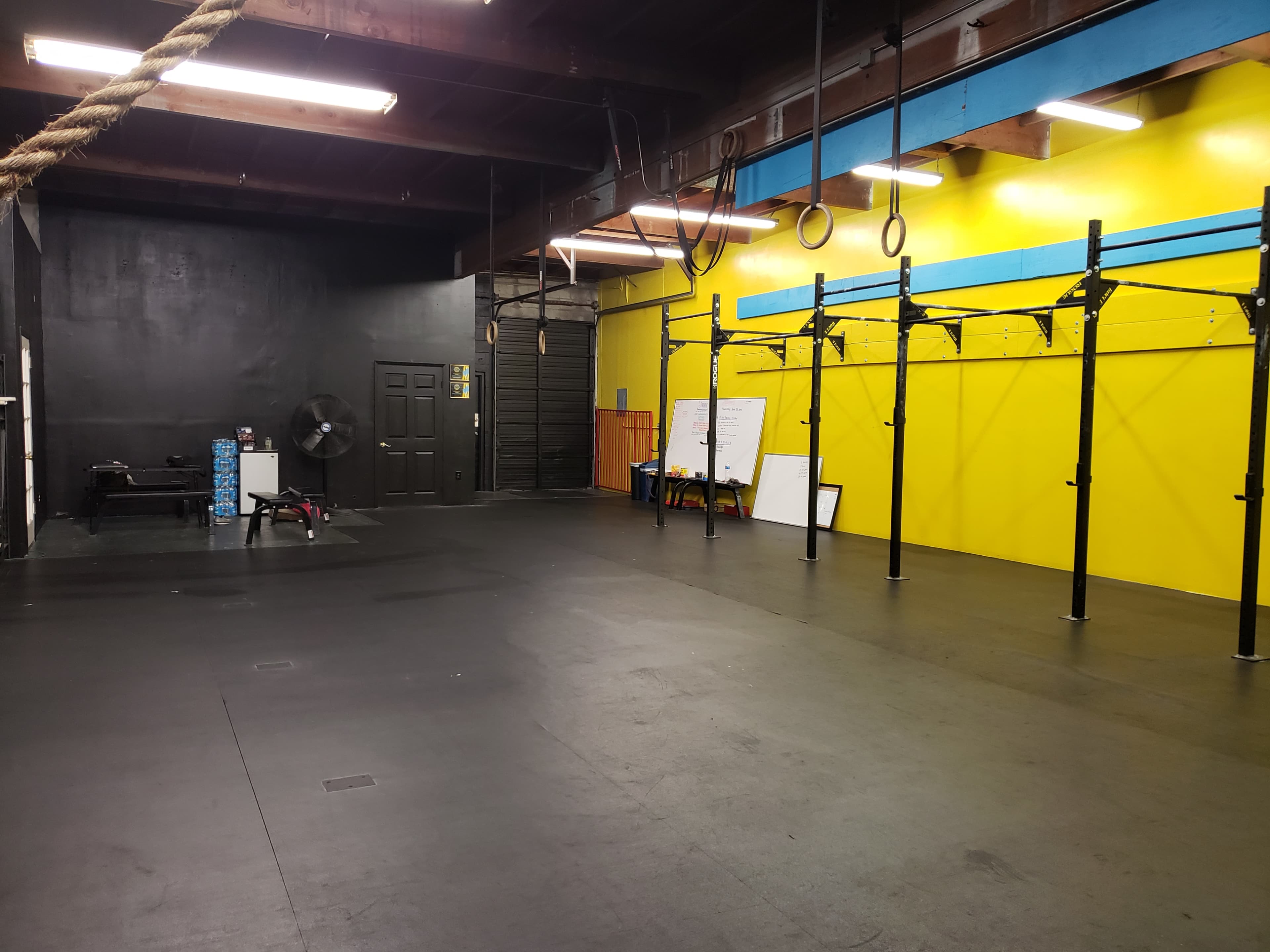 A spacious gym interior with a black and yellow color scheme, featuring workout equipment, pull-up bars, and a fan along one wall.
