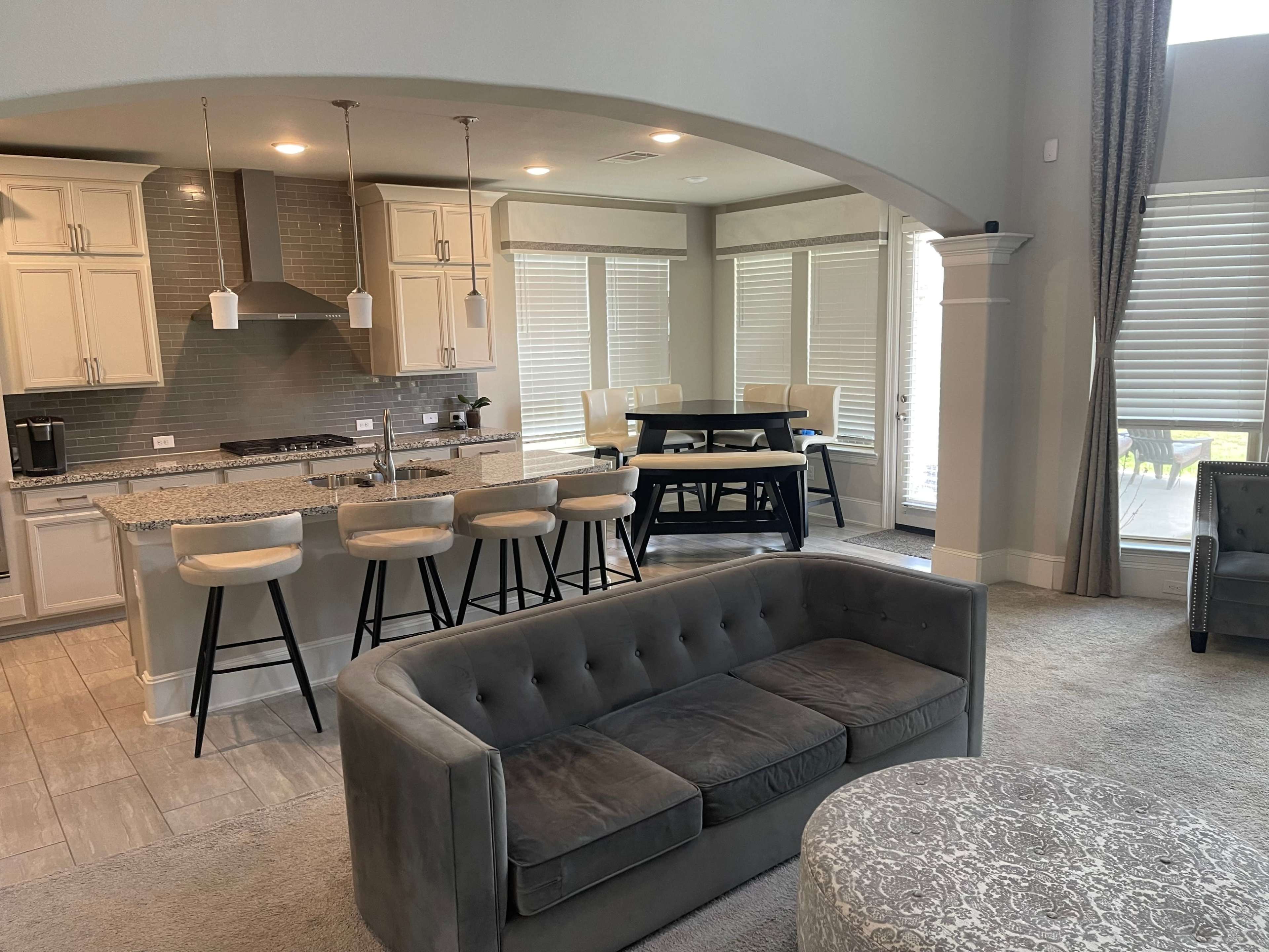 The image shows a modern, open-concept living and dining area with a kitchen featuring a large island and seating, flanked by a gray sofa and a round coffee table.