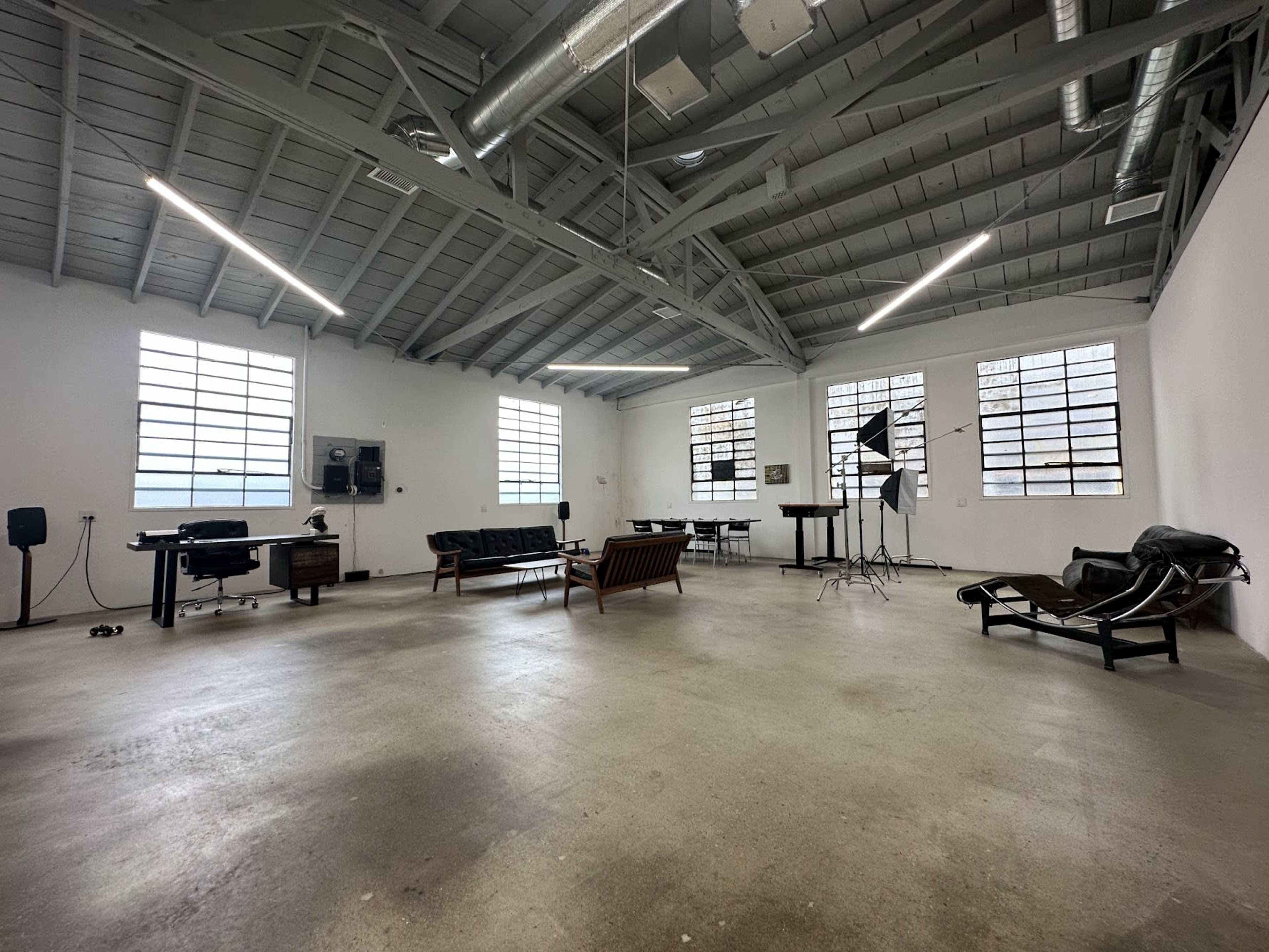 A spacious, industrial-style room features concrete floors, large windows, and minimal furniture including a couple of sofas, a table, and photography equipment.